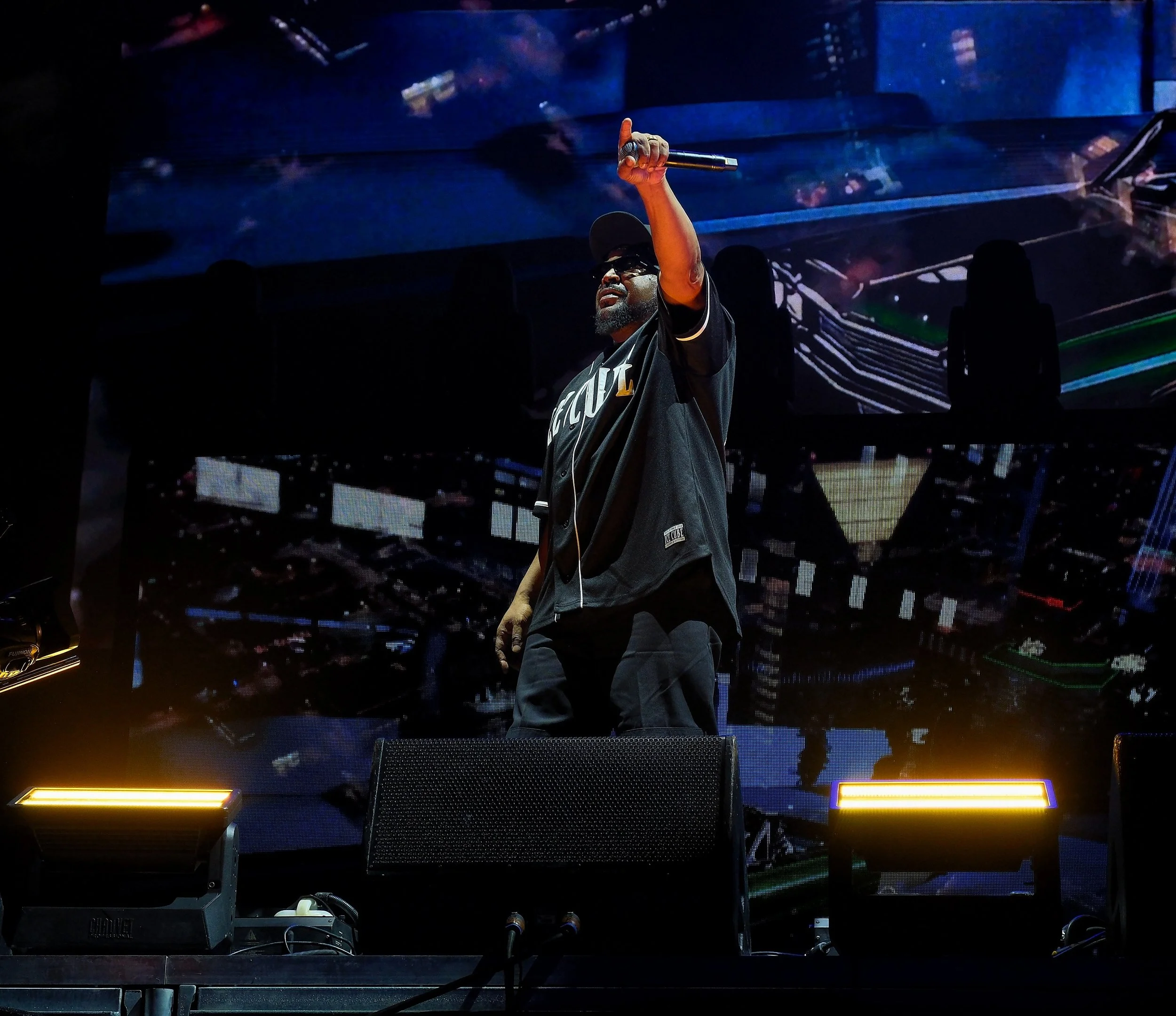 A performer on stage holding a microphone and raising one finger, with digital screens in the background displaying abstract images.