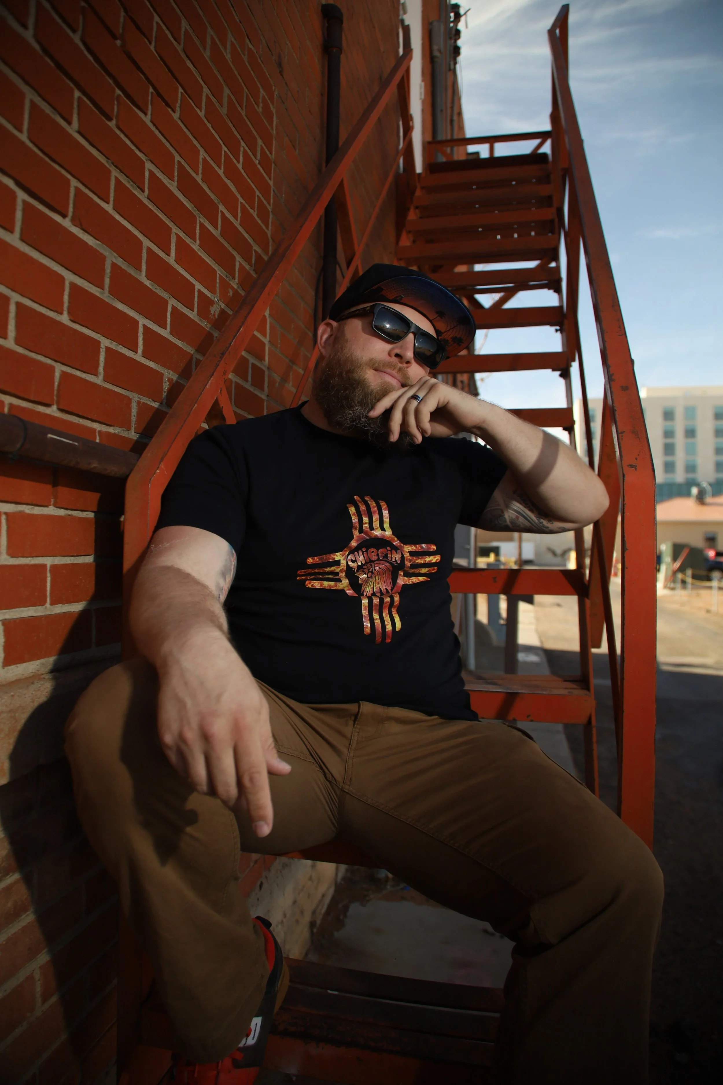 A man with a beard wearing sunglasses, a black hat, and a black T-shirt sitting against a brick wall with orange metal stairs behind him.