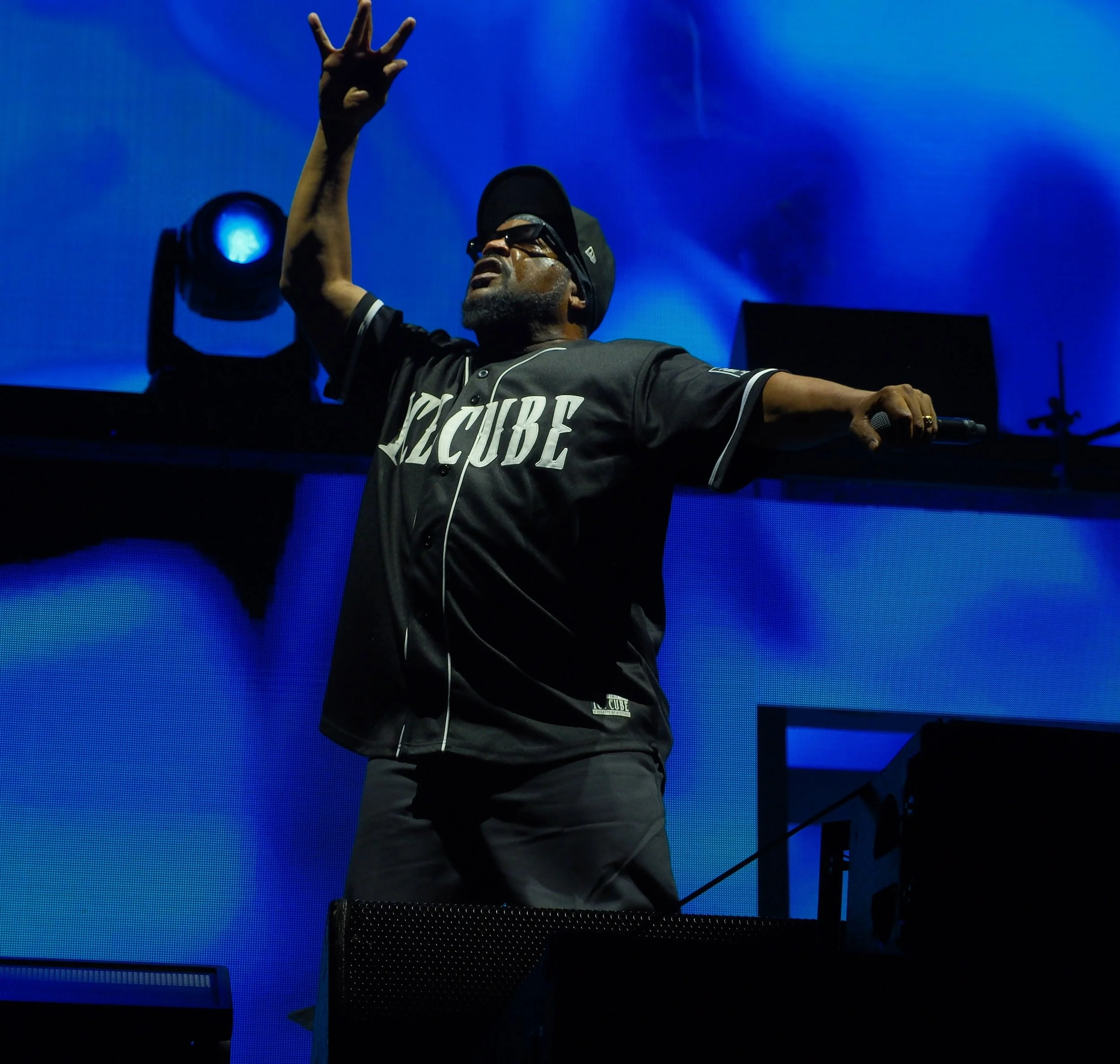 A DJ wearing sunglasses and a black cap is performing on stage with a blue backdrop. The DJ is dressed in a black shirt with white lettering and is reaching up with one hand. There are stage lights and equipment visible.