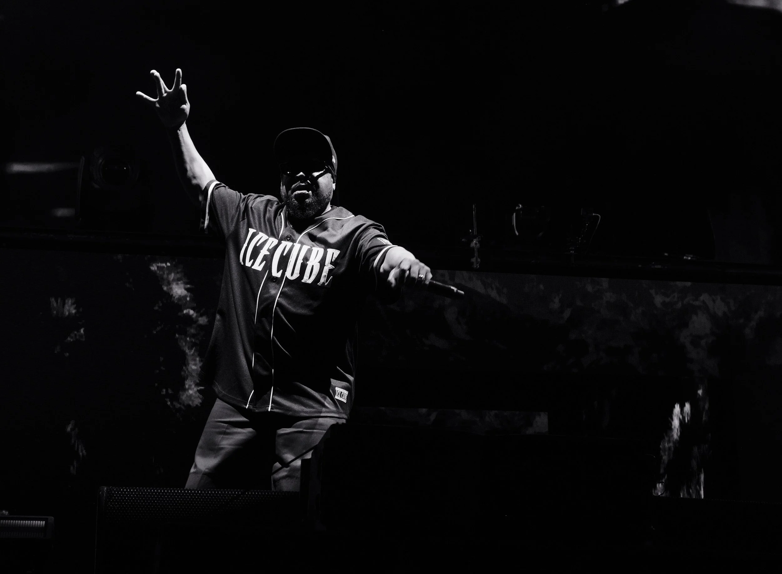 A man wearing sunglasses and a cap, dressed in a jacket with 'ICE CUBE' written on it, performing on stage with one arm raised.