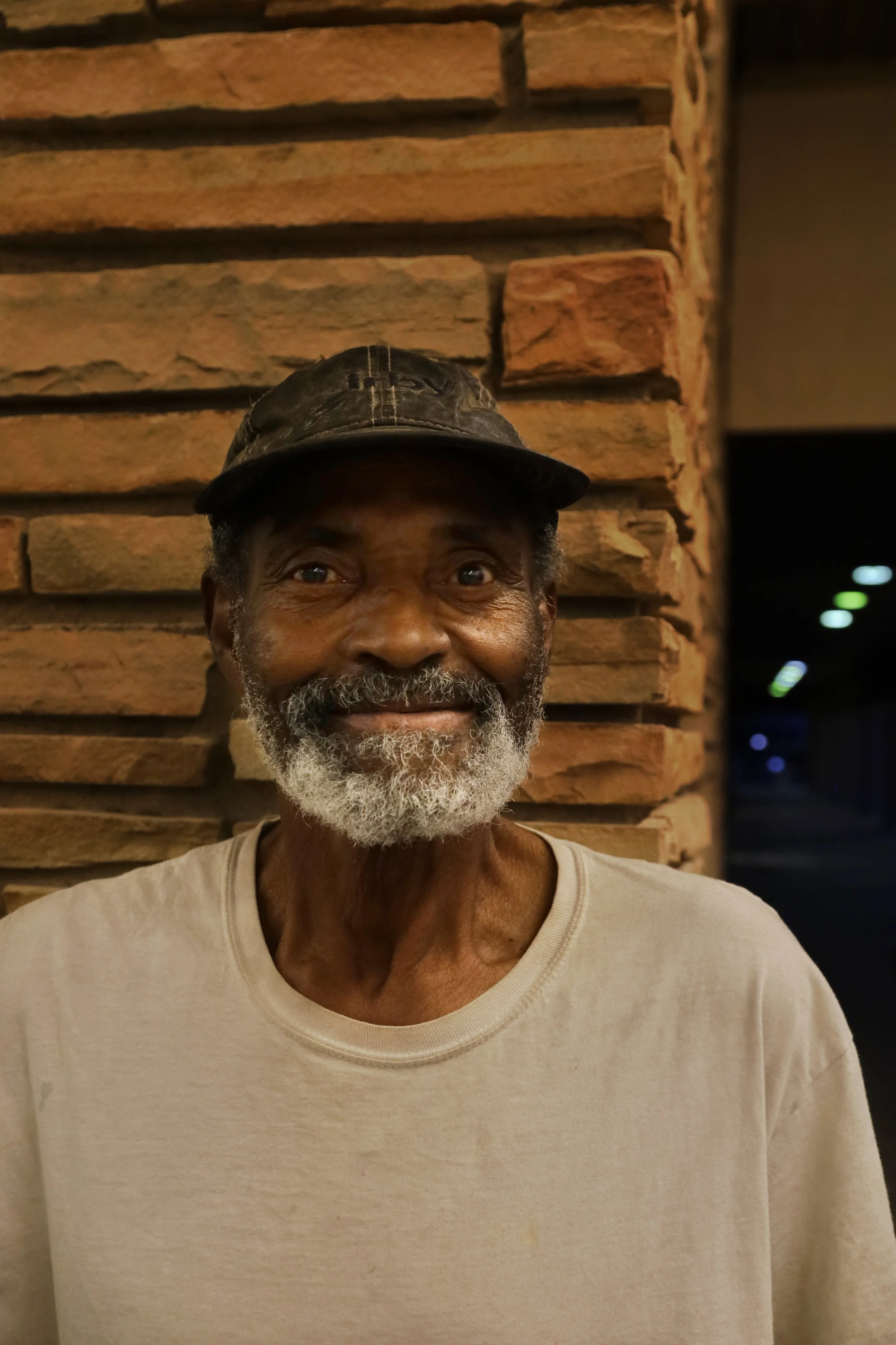 A smiling elderly man with a gray beard and mustache wearing a black cap and a beige shirt, standing in front of a textured brick wall in an indoor setting.