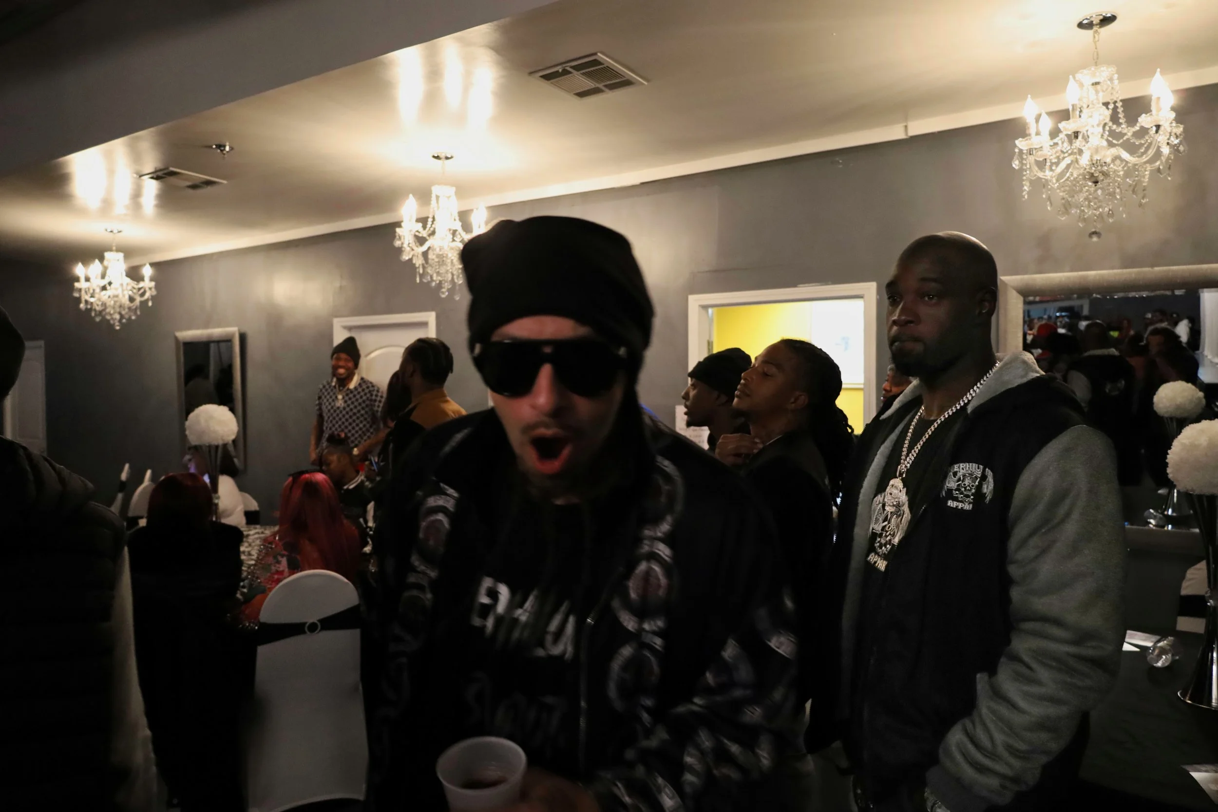 Group of people at a party in a decorated room with chandeliers, including two men in the foreground and others in the background.