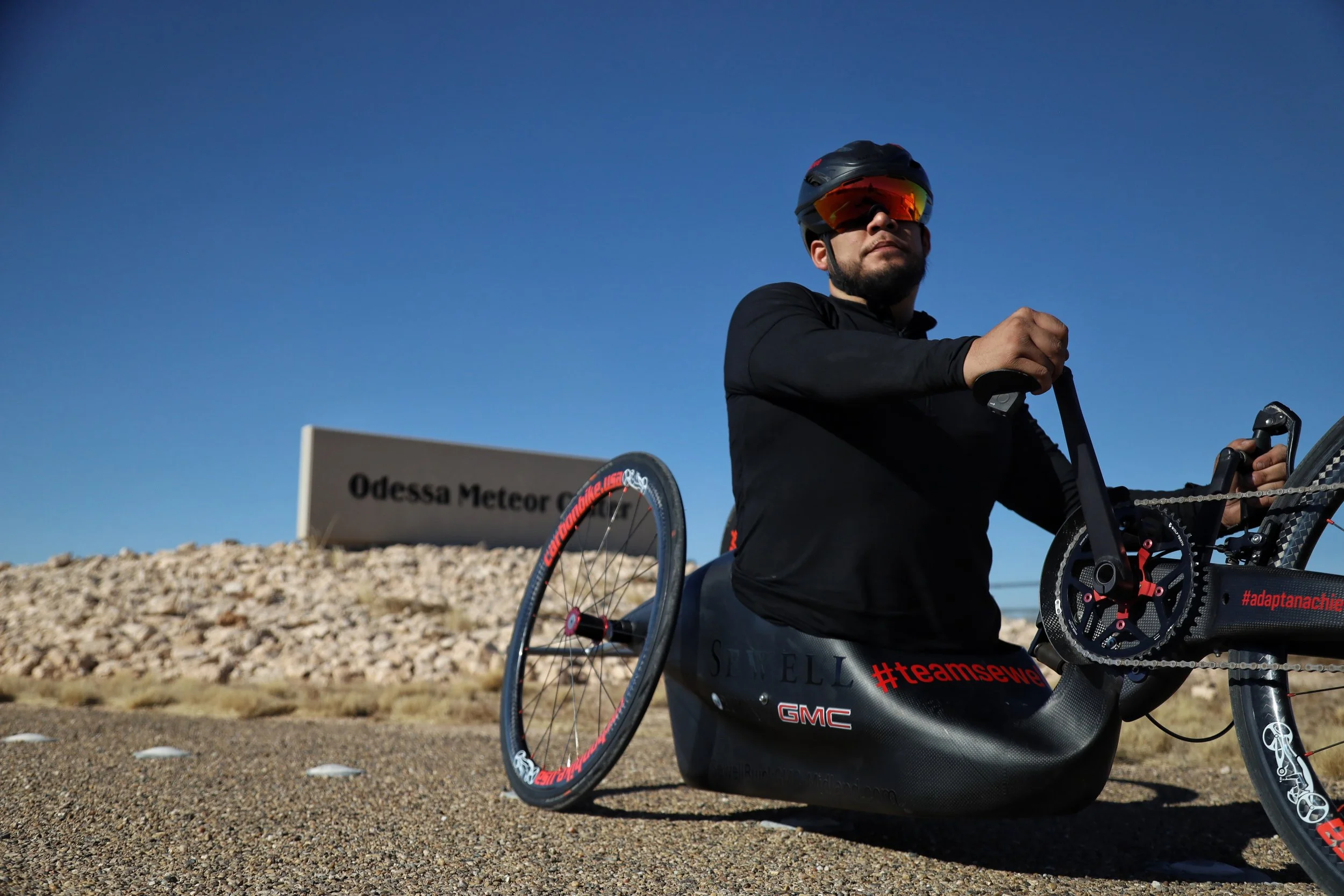 A person wearing a helmet and sunglasses sitting on a black racing wheelchair designed for high-speed track racing, with a sign in the background that reads 'Odessa Meteor.'