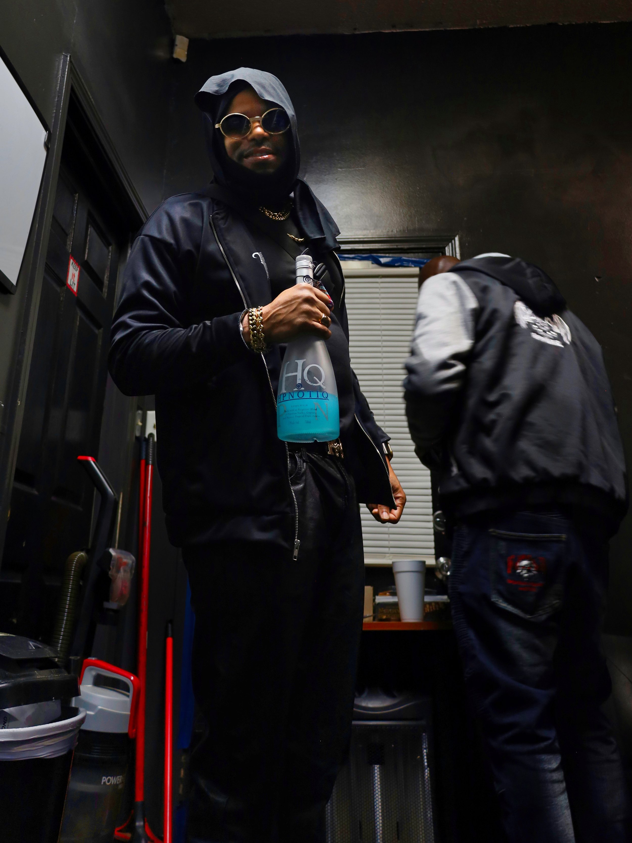 Person wearing sunglasses and a hooded jacket holding a spray bottle, standing indoors with another person in the background.
