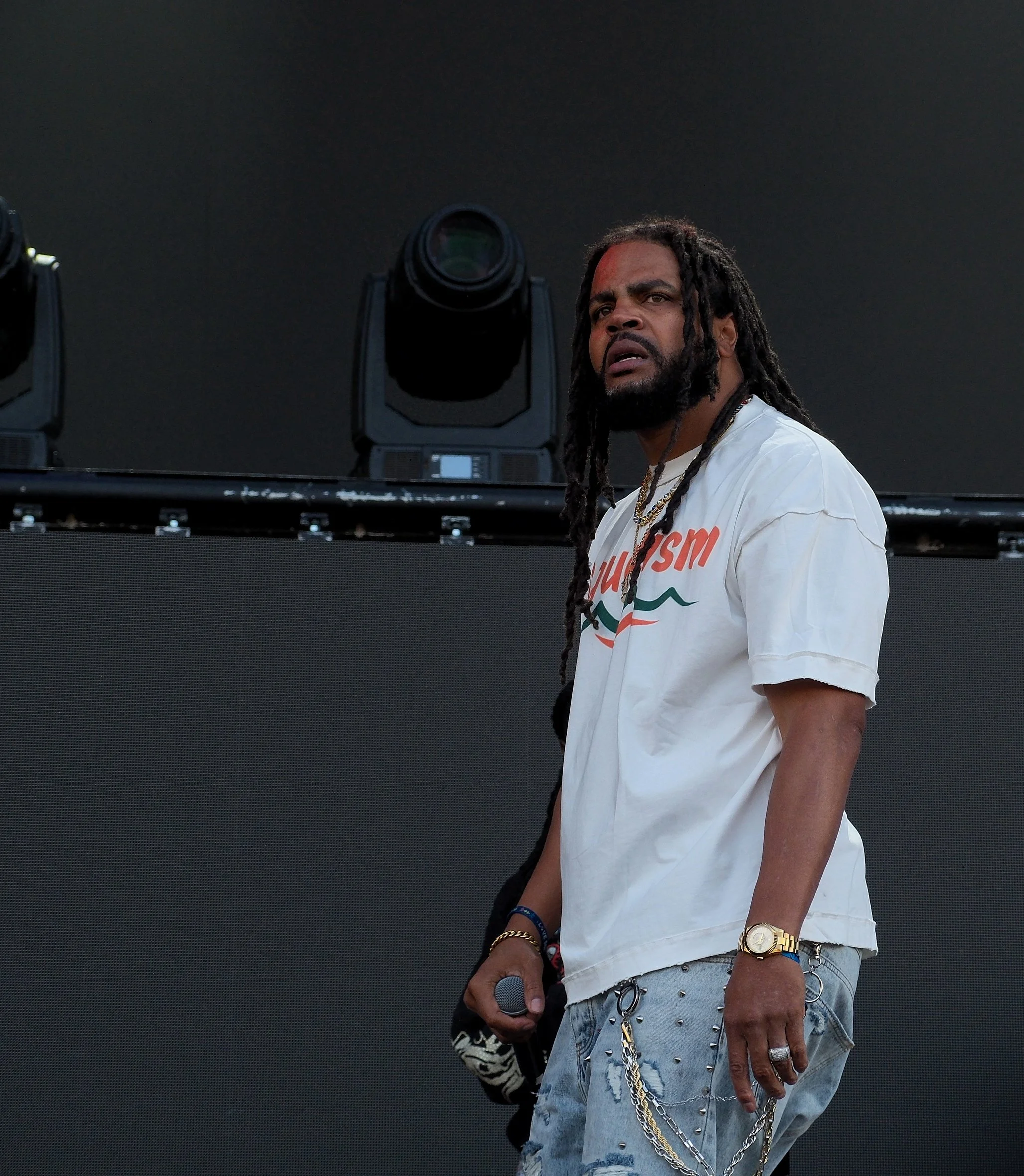 A man with dreadlocks and a beard stands in front of a dark background, wearing a white t-shirt with orange and green lettering, layered with jewelry and accessories.