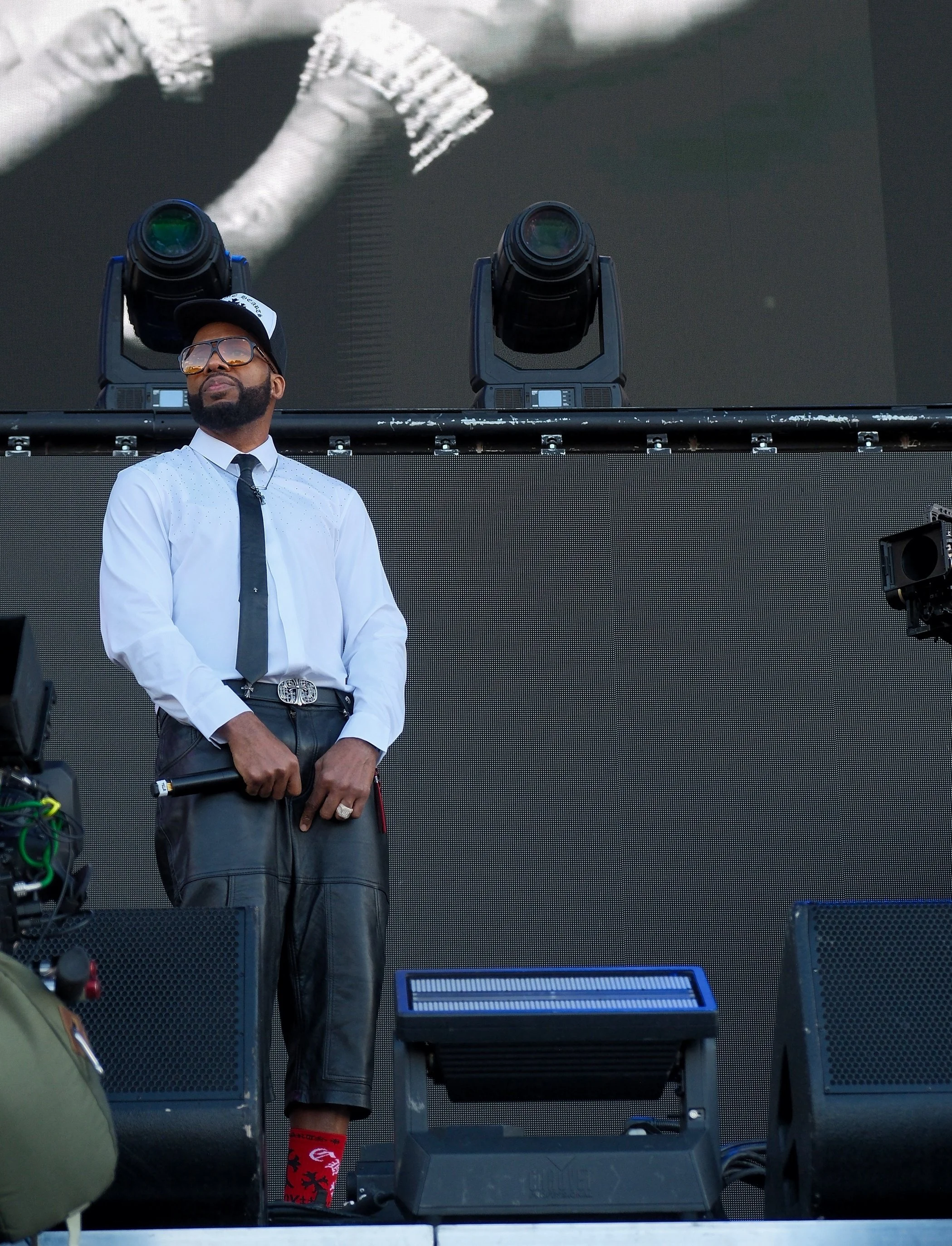 A man standing on stage during a performance, wearing a white shirt with small dots, black tie, black leather shorts, a black and white cap, sunglasses, and bright red socks with black patterns, behind professional stage equipment including laser pro