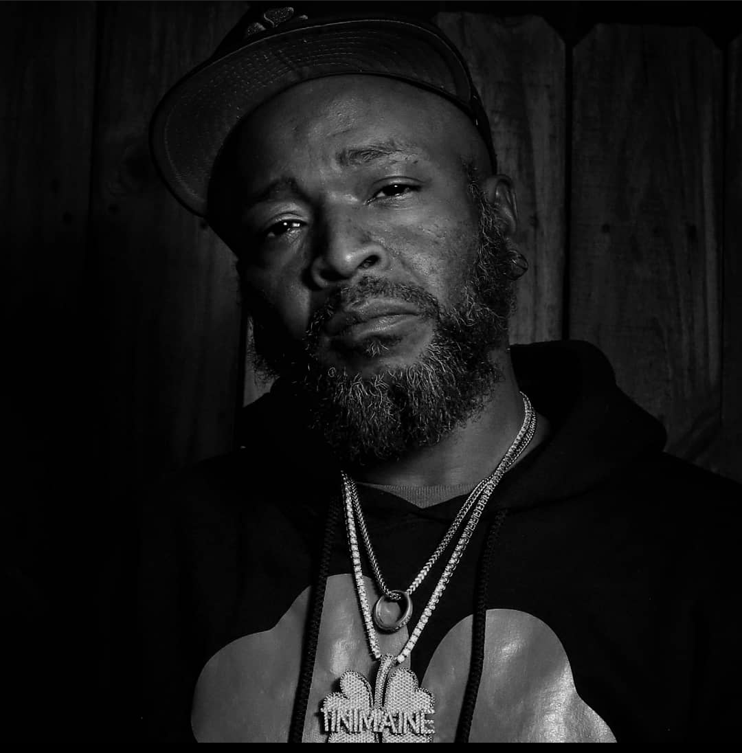 Black and white portrait of a man with a beard wearing a baseball cap, multiple necklaces, and a hoodie with a graphic design.