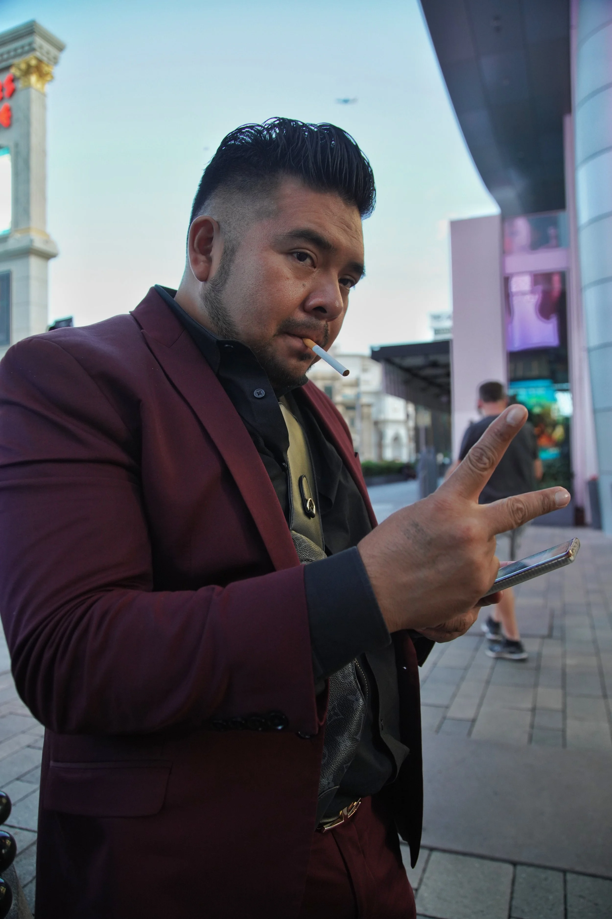 A man wearing a burgundy suit and black shirt, holding a smartphone in one hand, making a peace sign with the other, and smoking a cigarette in an urban area with buildings and a crowd in the background.