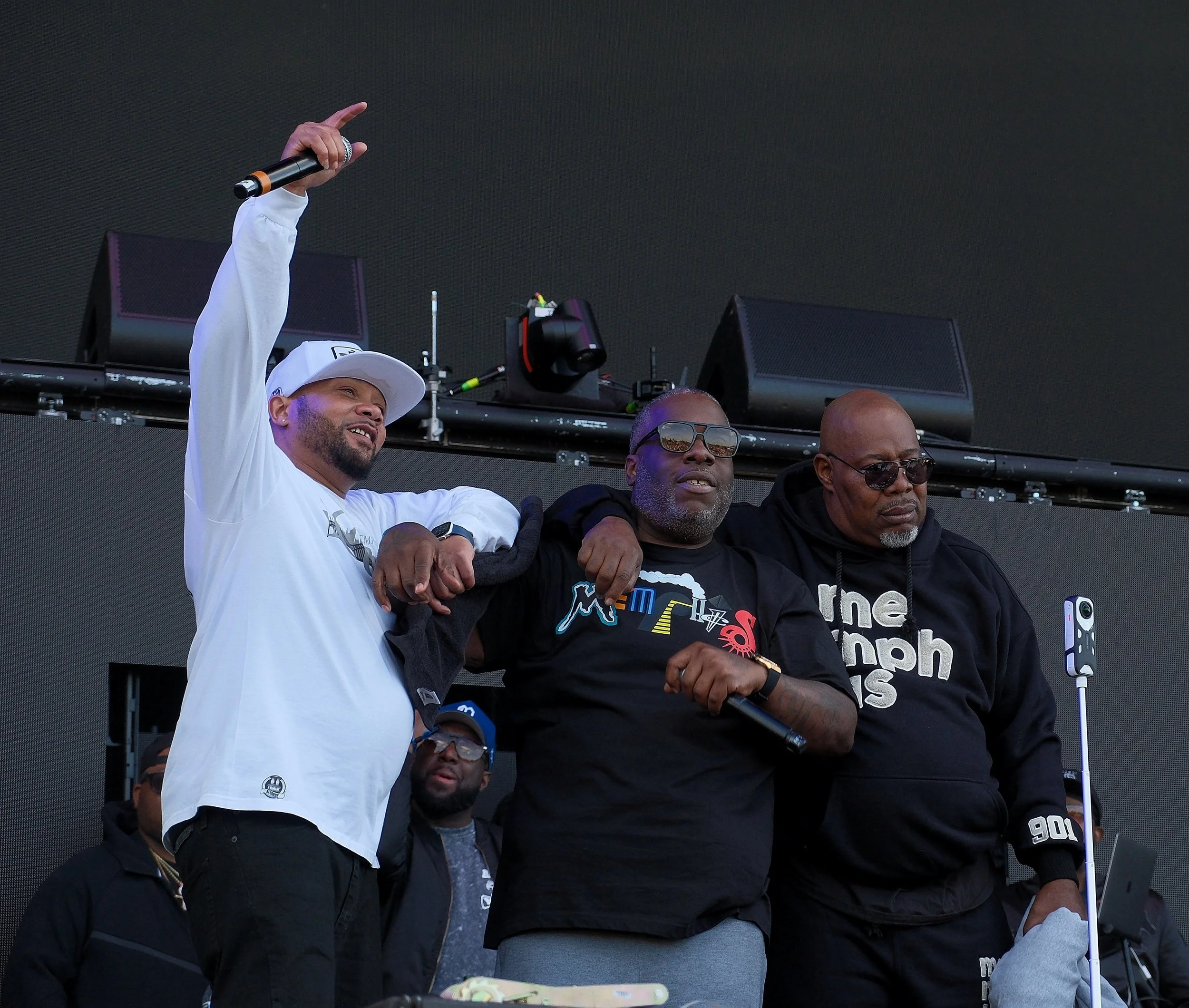 Group of people standing on stage, with one person holding a microphone and raising their hand, at an outdoor event or concert.