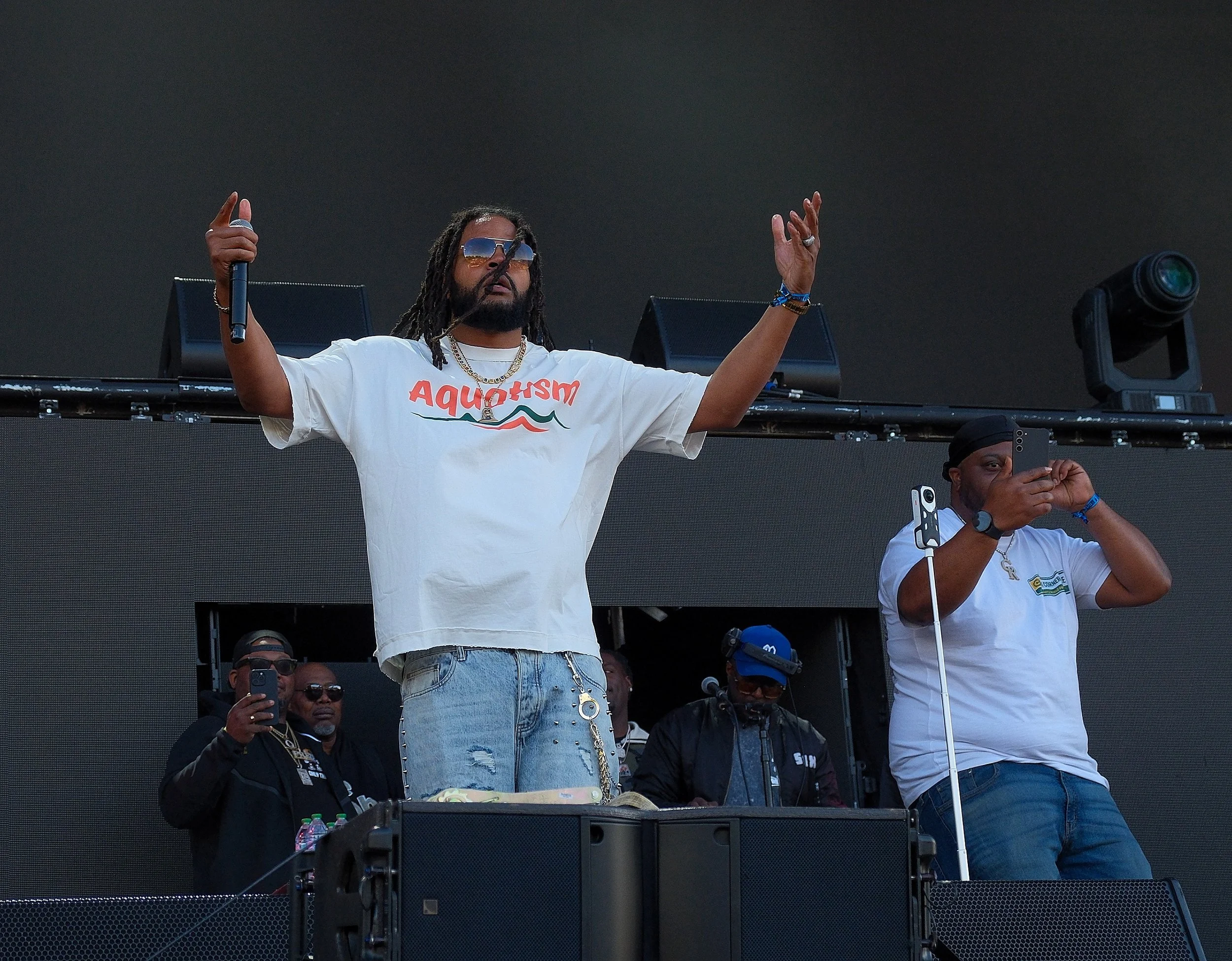 A man with long dreadlocks, sunglasses, and a white T-shirt stands on a stage with his arms raised, holding a microphone, surrounded by a crowd of people, some taking photos or videos, in an outdoor event.