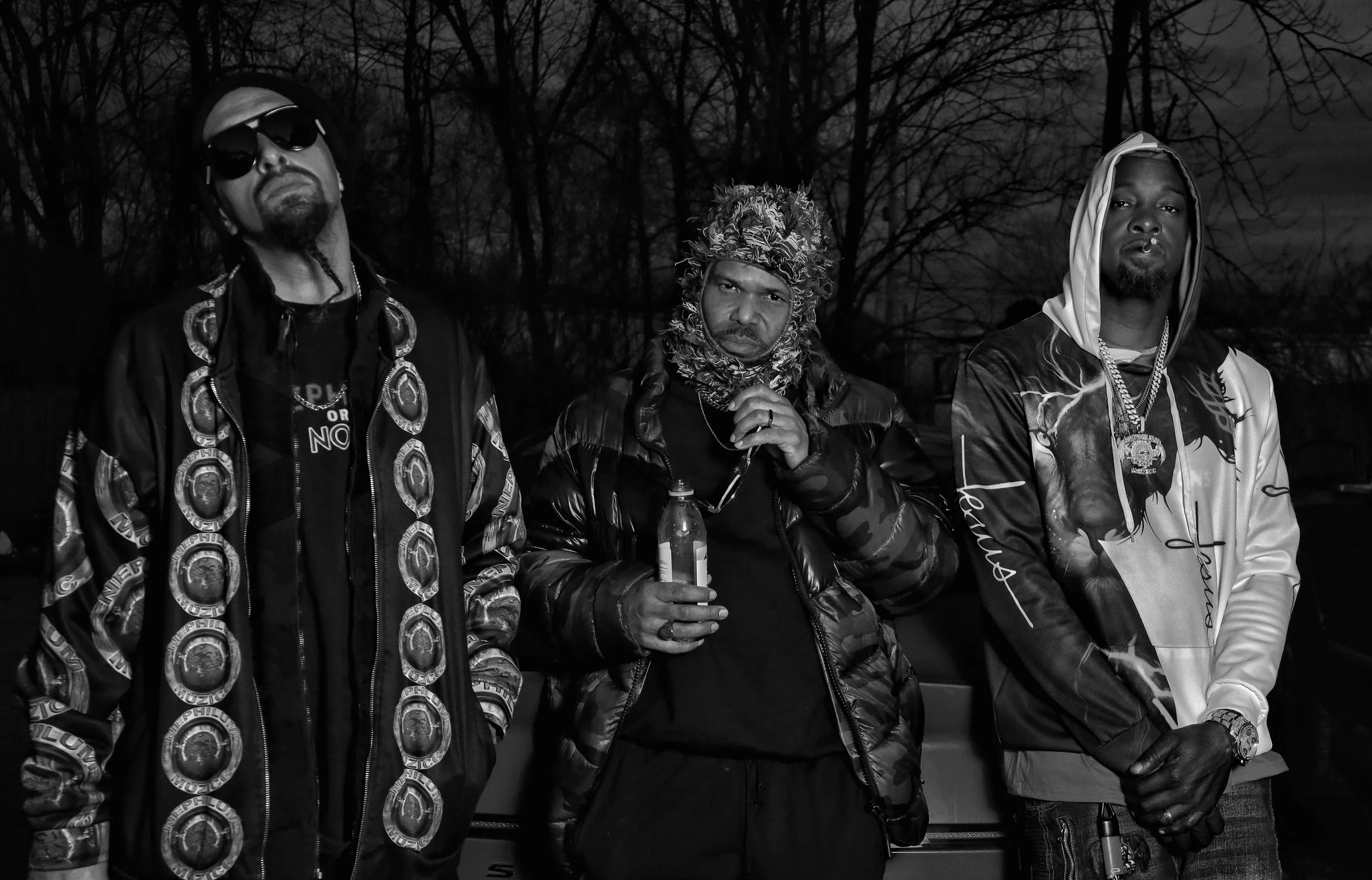 Group of three men standing outdoors in a wooded area at night, wearing stylish clothing and accessories, with serious expressions.