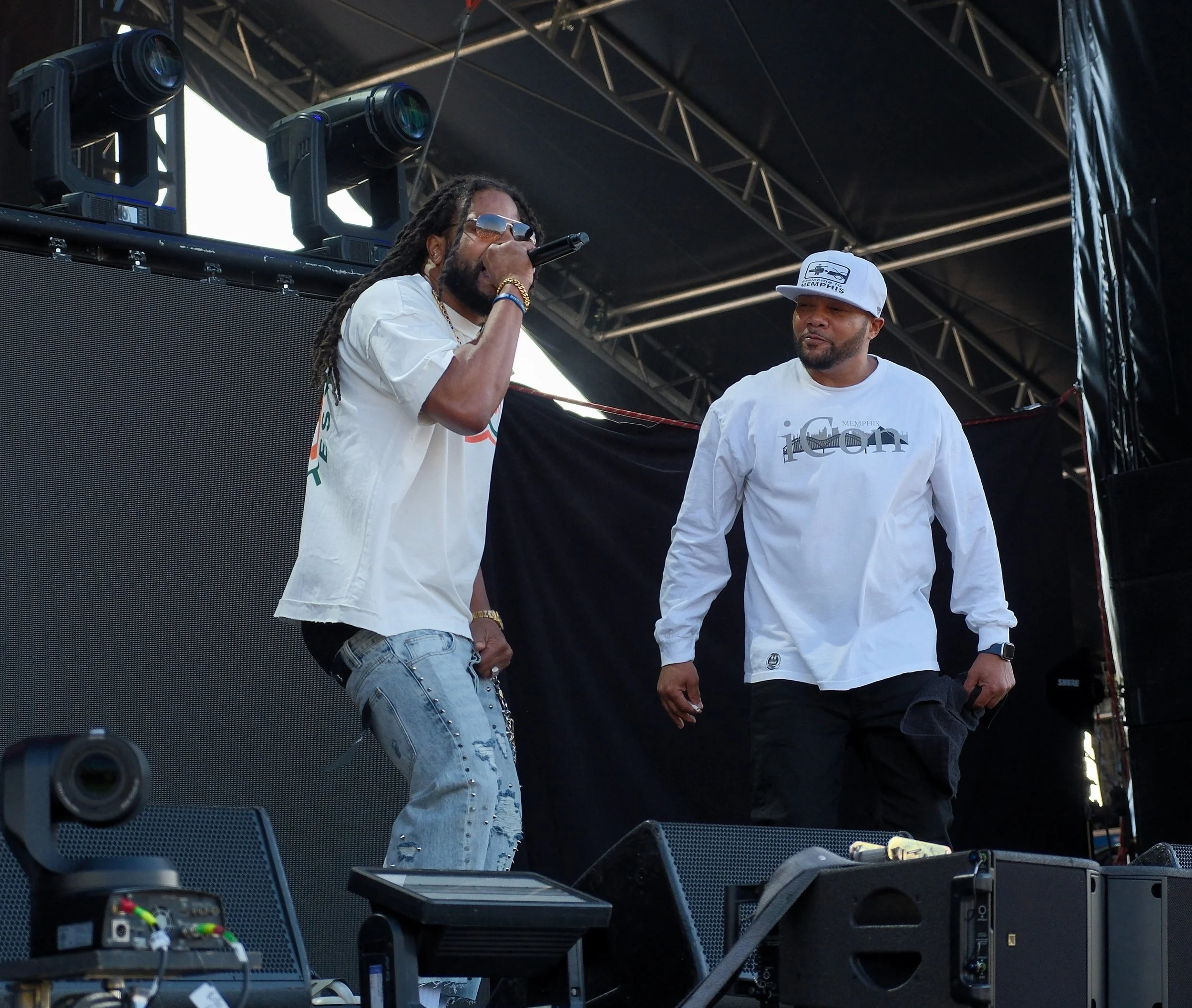 Two men are performing on a stage, one is speaking into a microphone and the other is standing nearby. They are dressed casually, and there are large stage lights and equipment surrounding them, with a black overhead canopy.