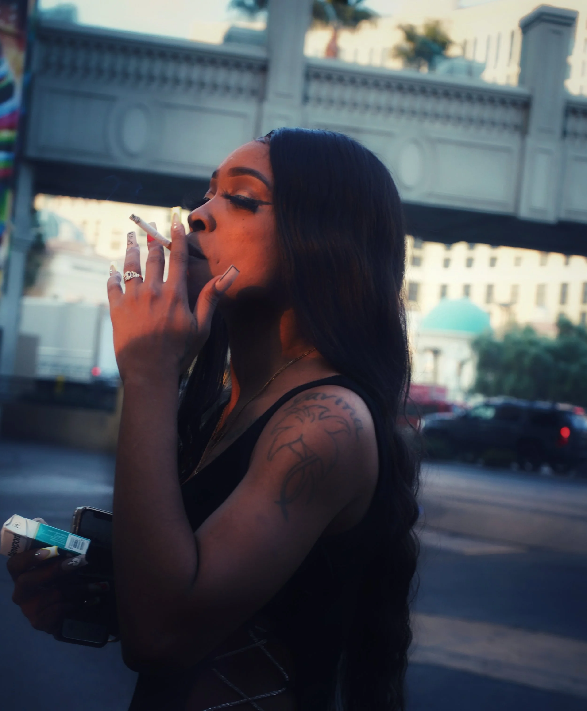 A woman with long black hair and tattoos on her arm is smoking a cigarette outdoors during the daytime, holding a pack and a lighter in her hands.