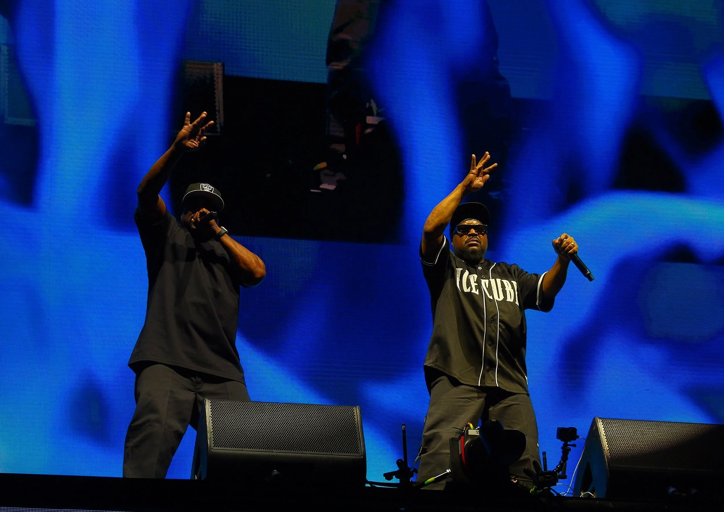 Two performers on stage, one holding a microphone and wearing sunglasses, the other raising their hand. Large blue screen in the background with a reflection of the stage and performers.