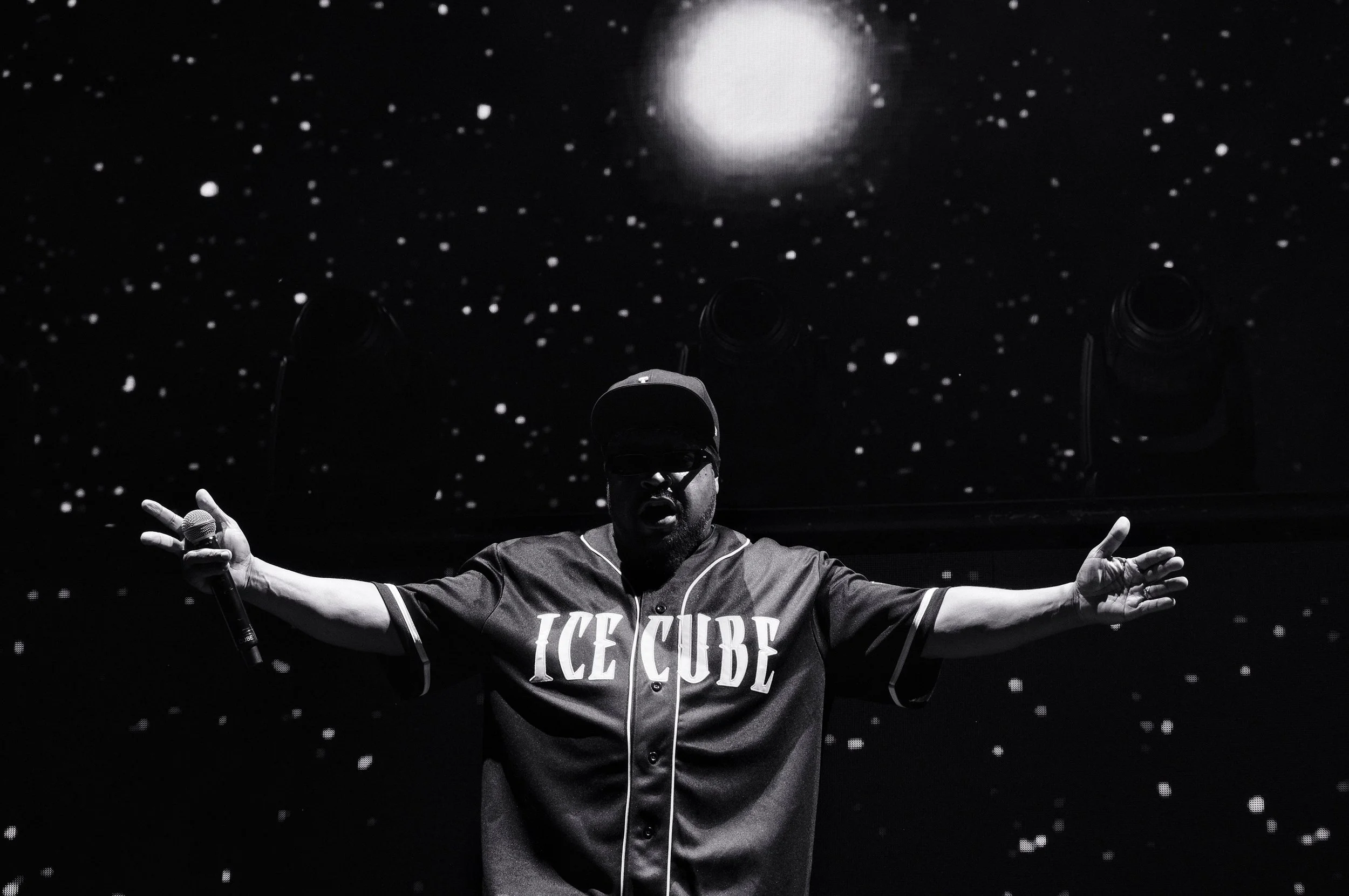 A performer on stage wearing a sports jersey that reads 'ICE CUBE', holding a microphone in one hand, with arms outstretched, under a starry background with a bright light overhead.