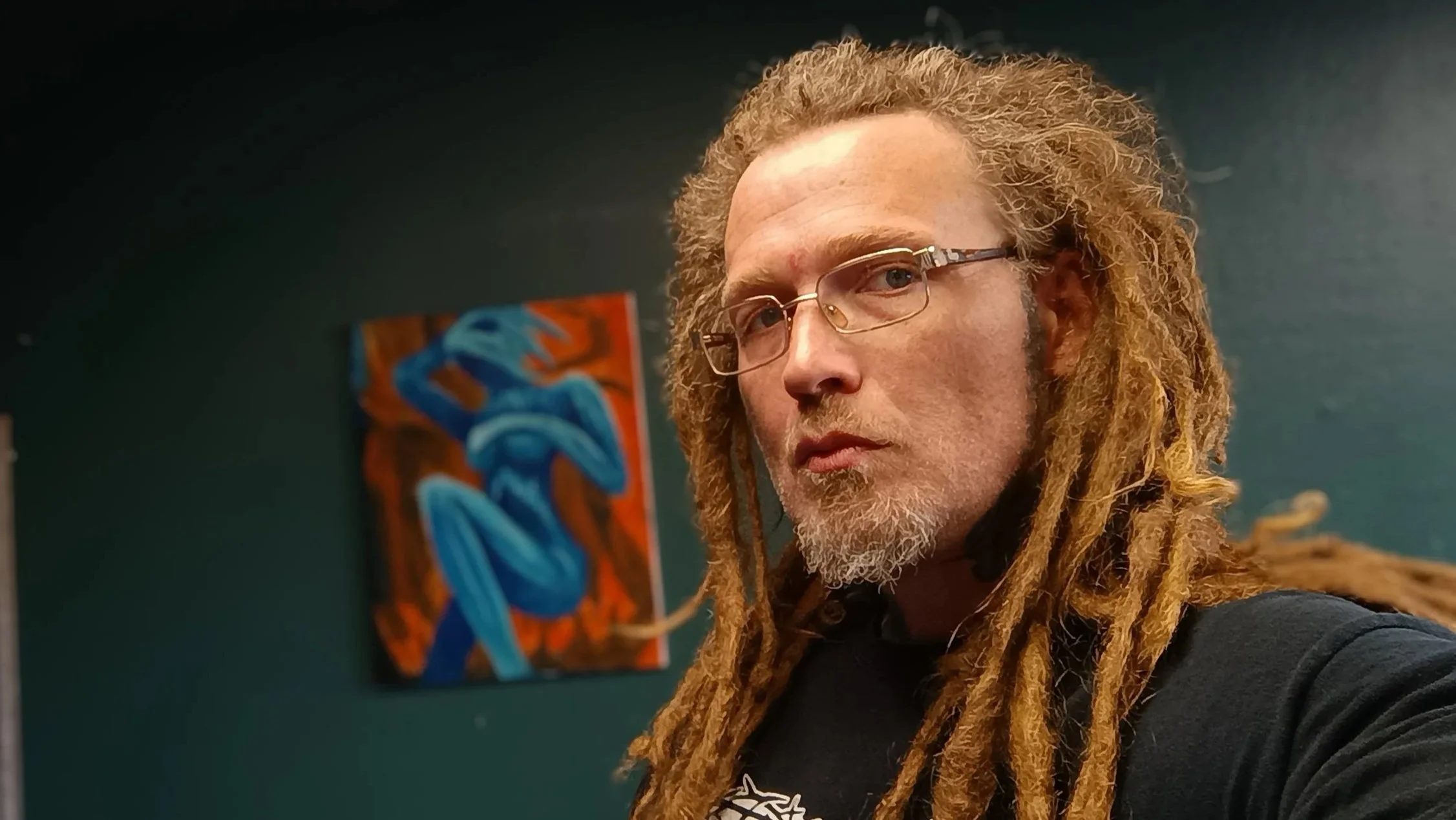 A man with light skin, long dreadlocks, glasses, and a goatee takes a selfie in front of a dark wall with a colorful abstract painting featuring blue leaves.