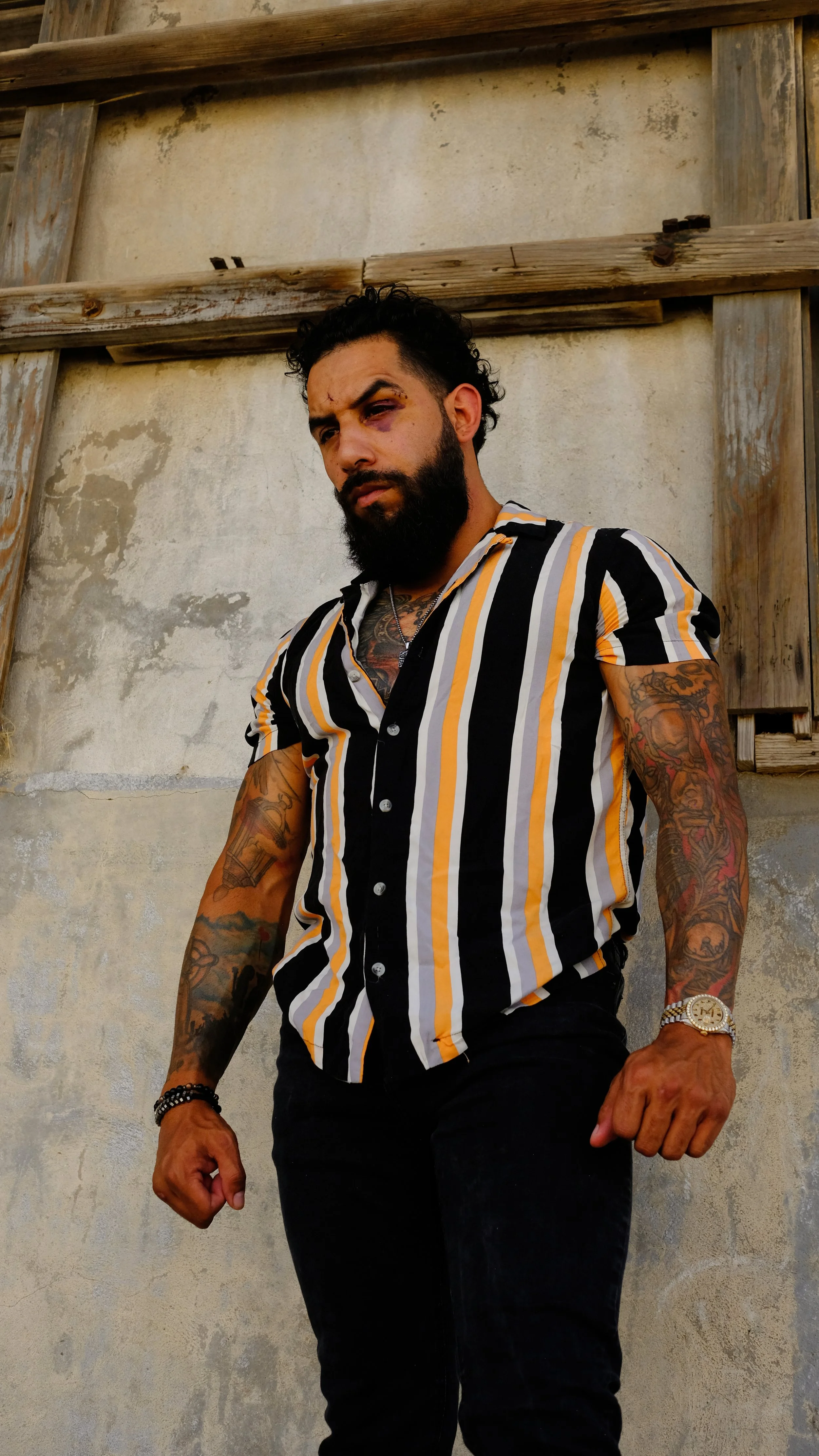 A man with a beard and tattoos wearing a black, white, and yellow striped short-sleeve shirt, standing against a weathered wall with wooden beams.