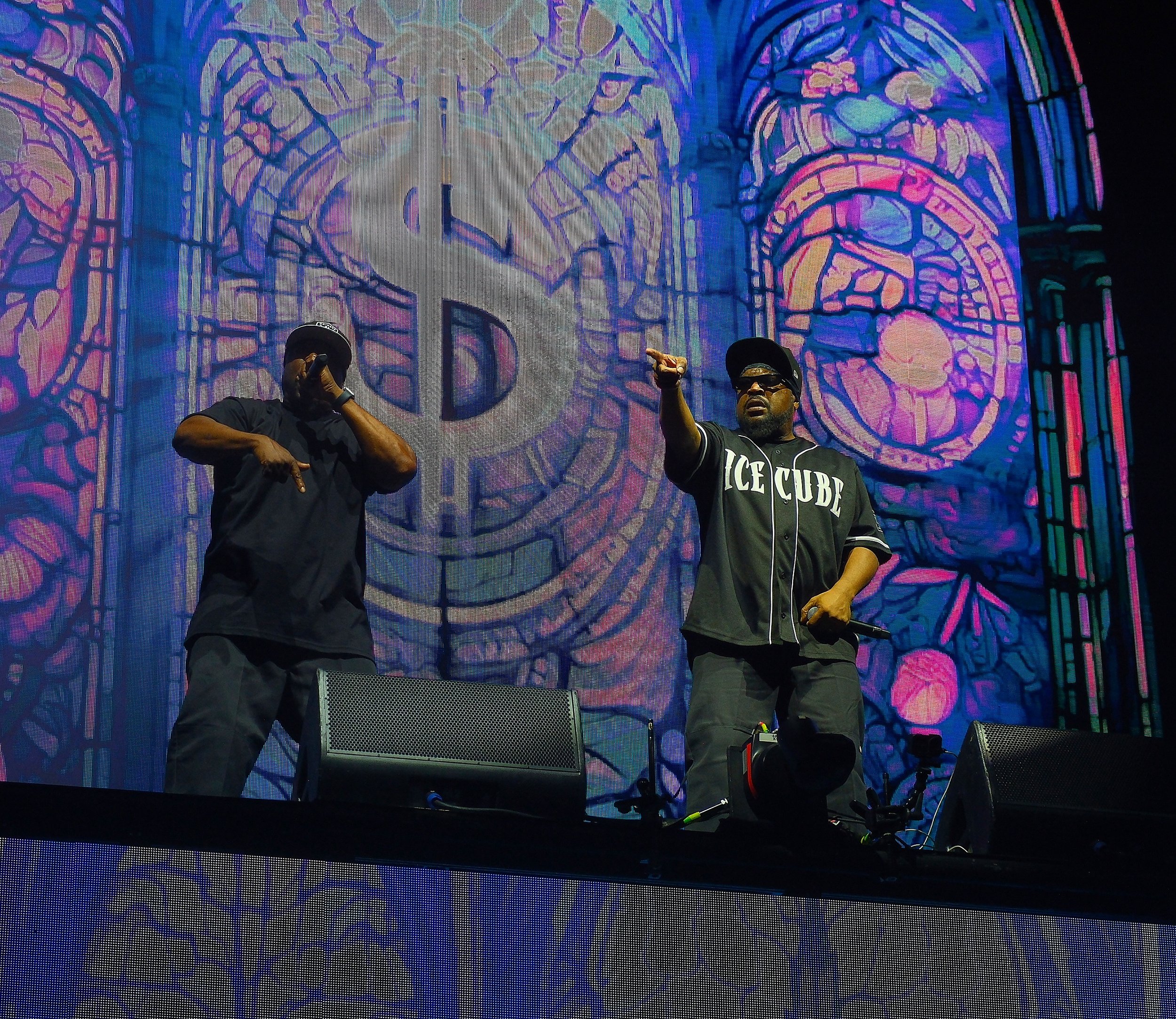 Two men, one in a black shirt and the other in a black Oklahoma City Thunder jersey, wearing sunglasses and black caps, performing on stage with a large colorful stained glass window projection in the background.