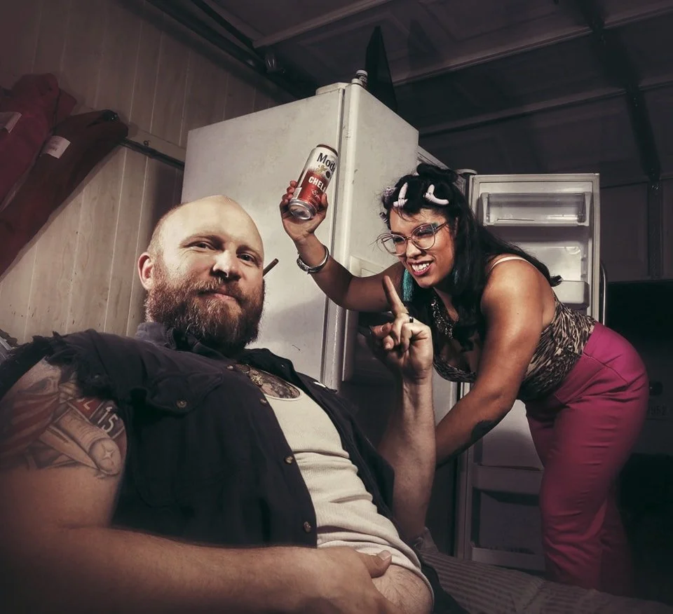A man with a beard and tattoos sitting in front of an open refrigerator, while a woman with glasses, hair rollers, and vibrant clothing leans over, holding a beer can and making a playful gesture.