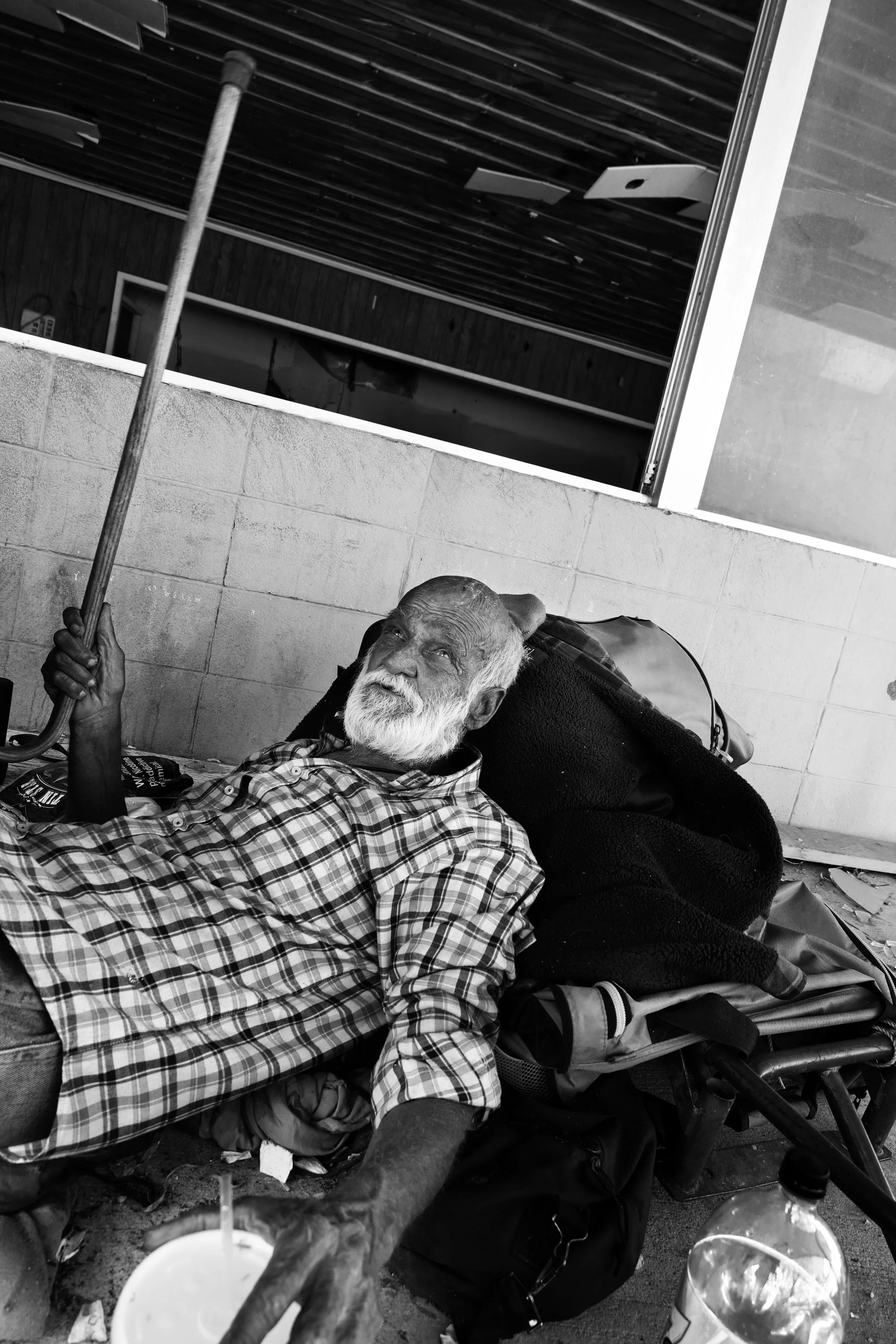 An elderly man with a white beard lying on a bed outdoors, holding a large umbrella handle, with a cup and bottled water nearby, against a brick wall and industrial background.