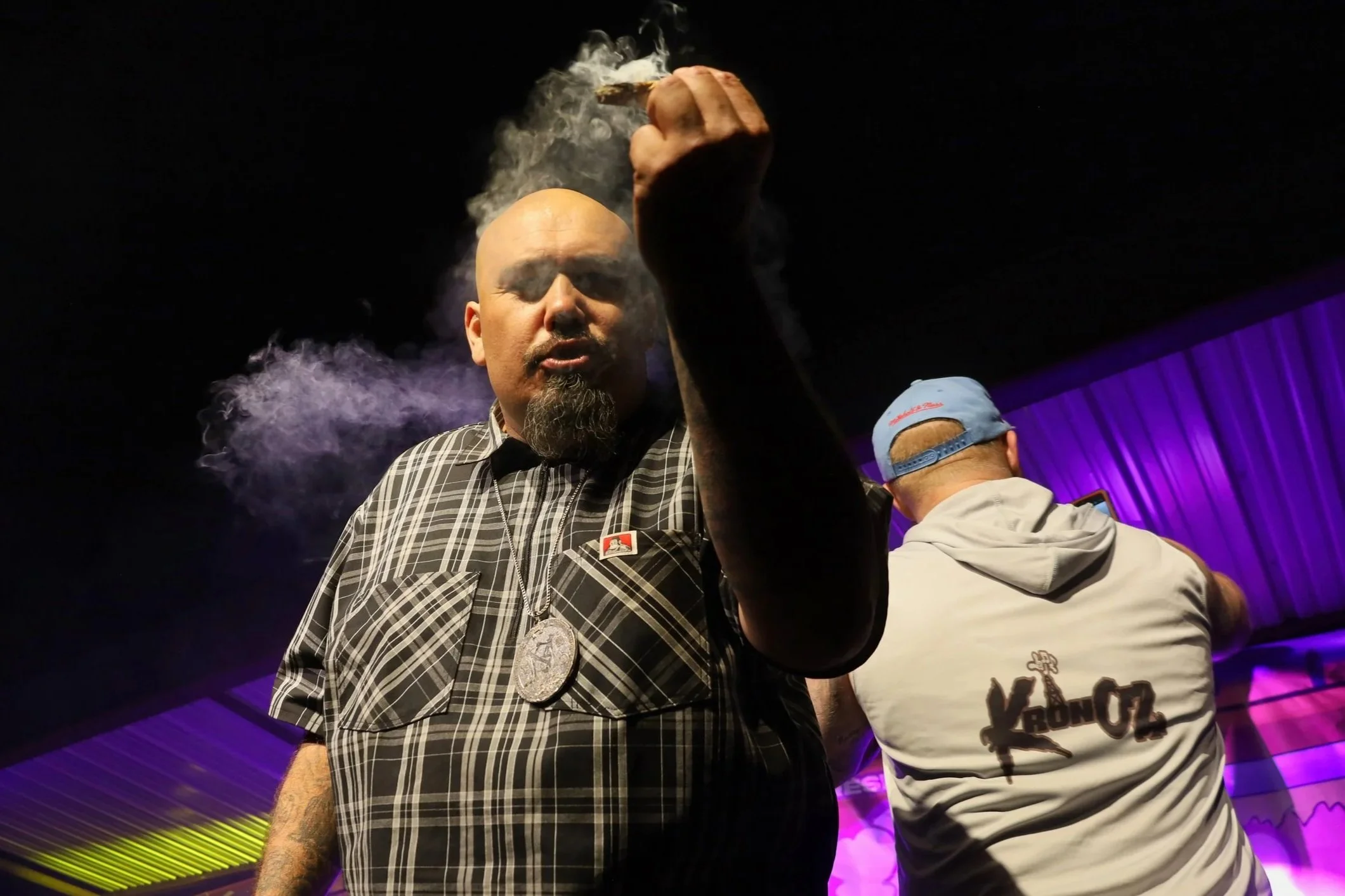 A bald man with a goatee, wearing a plaid button-up shirt and a large medallion necklace, appears to be vaping as smoke surrounds him, with purple lighting and another person in a gray hoodie and cap in the background.