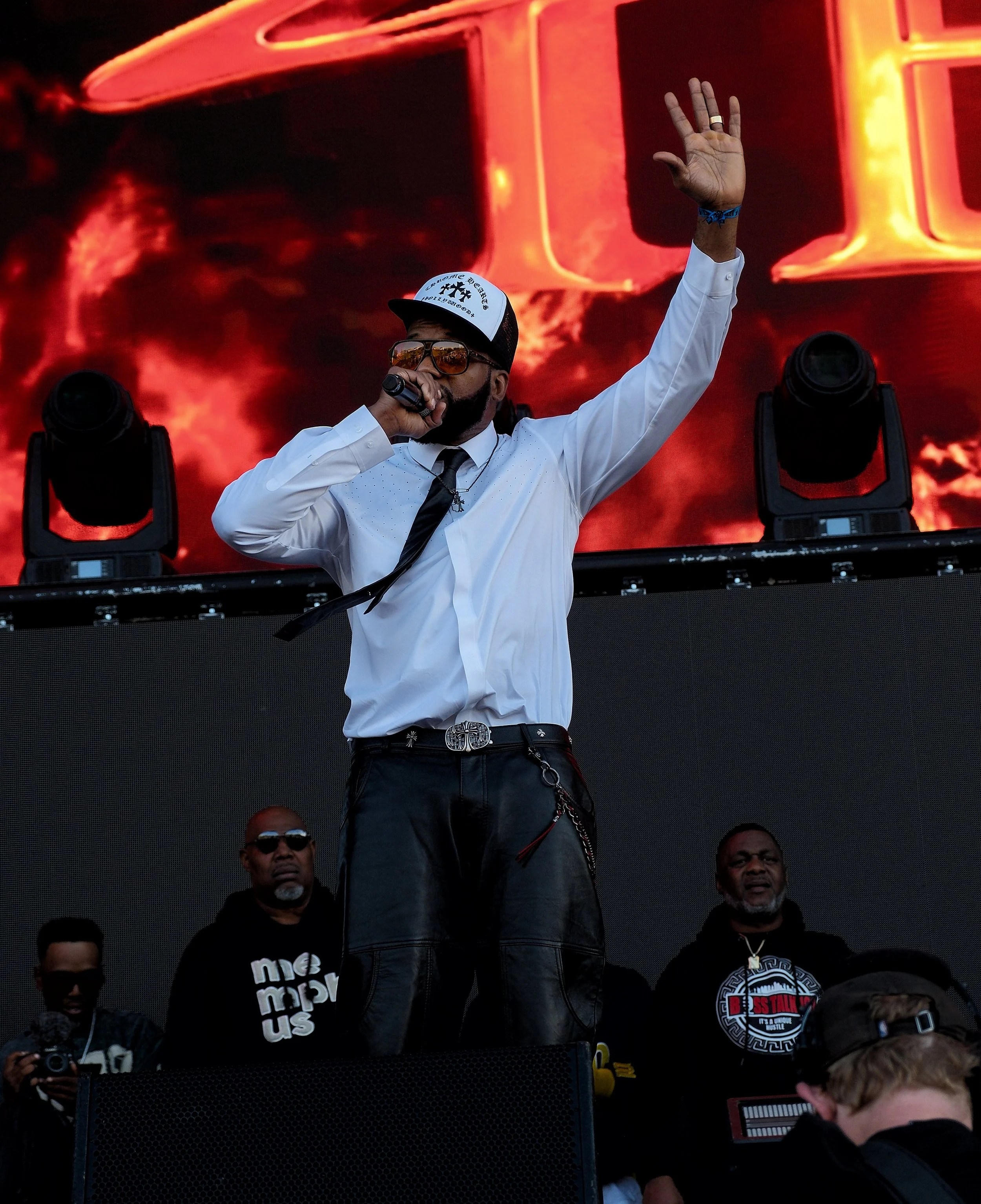 A man in sunglasses, a white dress shirt, black pants, and a baseball cap with a message, holding a microphone and raising his hand, standing on a stage with flashing lights and a large LED screen behind him. Several other people are around him.