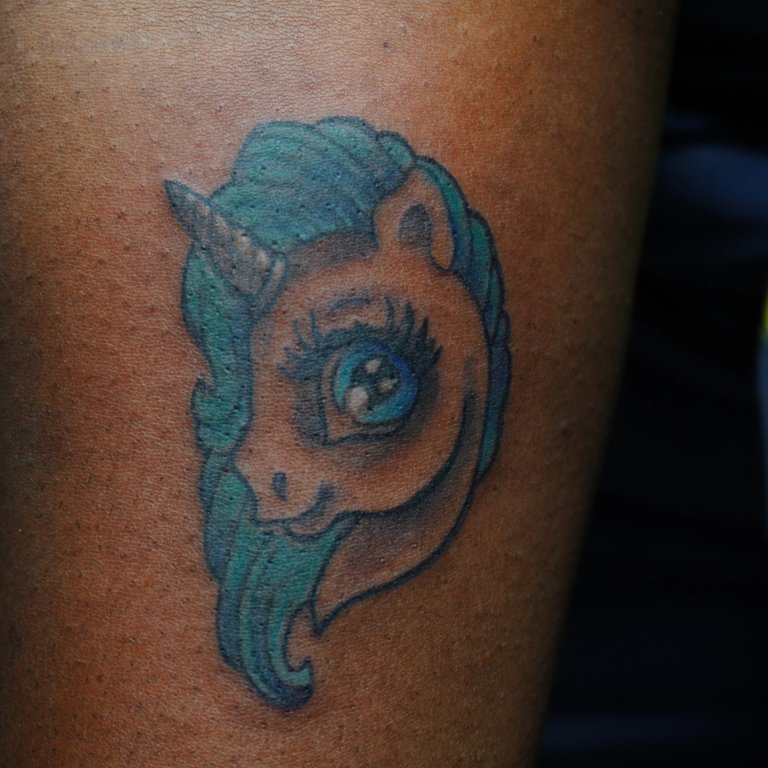 Tattoo of a unicorn with a blue mane, big eye, and a horn on the head.