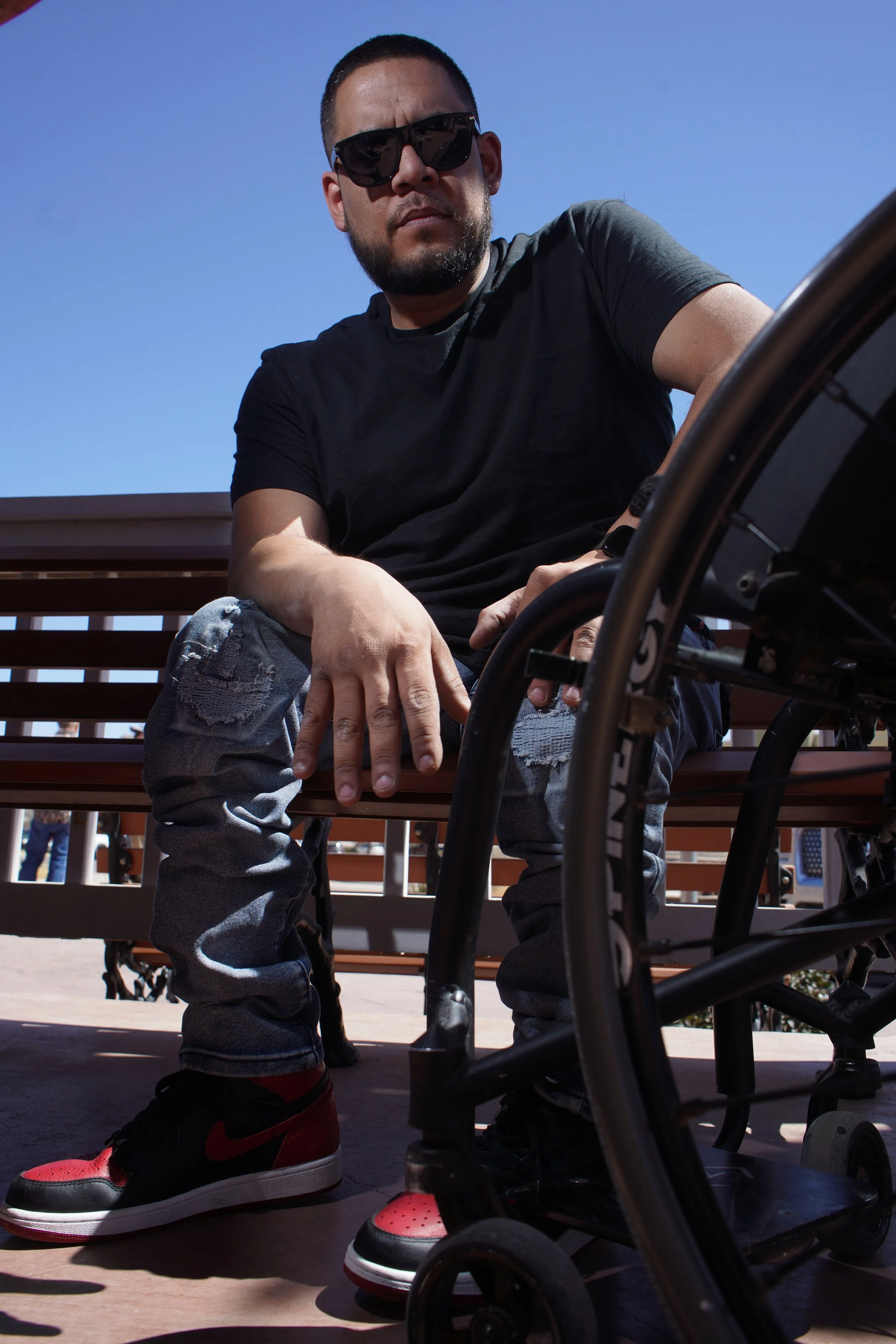 Man sitting on a park bench wearing sunglasses, a black T-shirt, ripped jeans, and red/black sneakers, with a wheelchair in front of him under a clear blue sky.