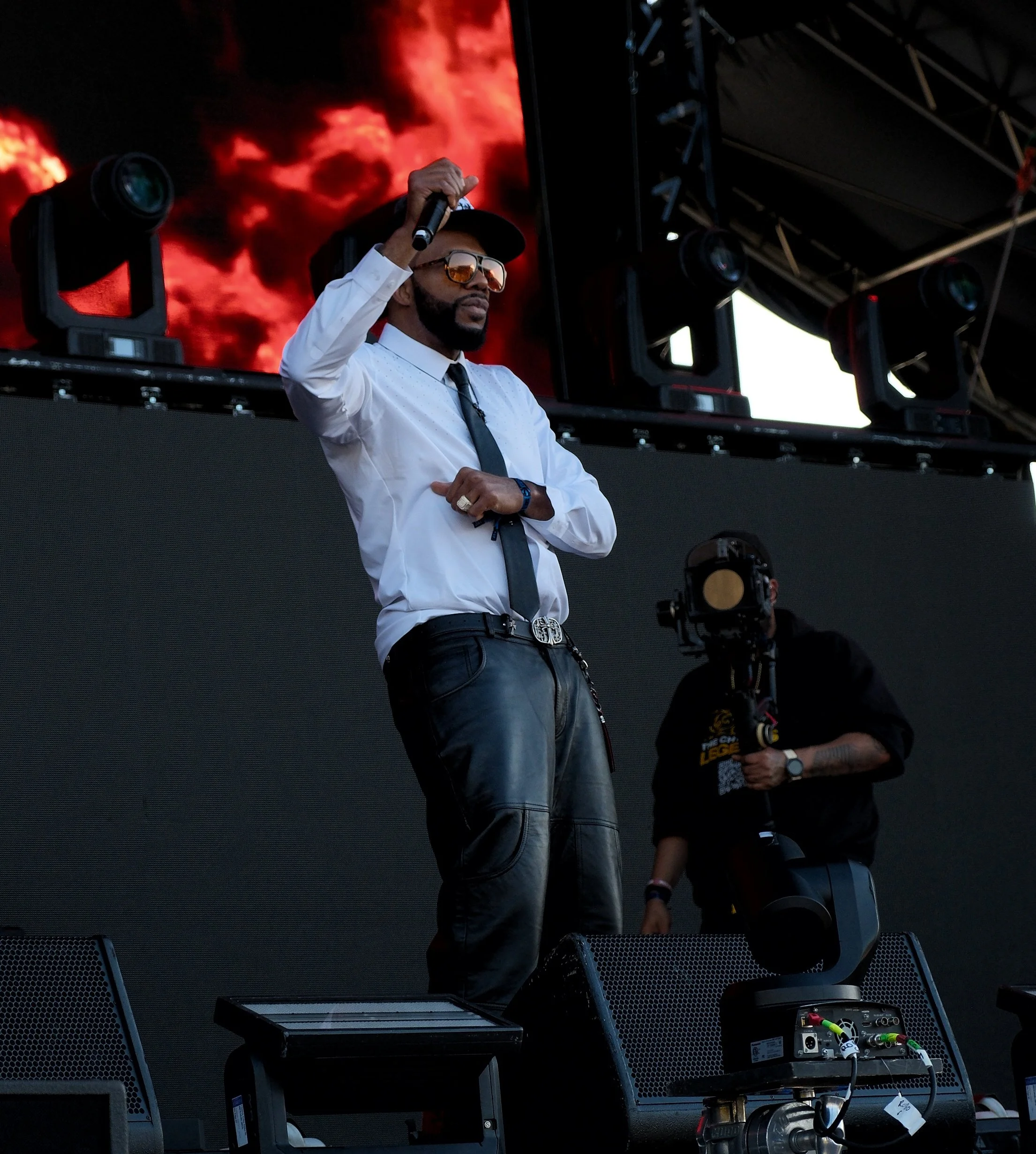 A man wearing sunglasses, a white shirt, and a black tie standing on a stage with stage lighting and equipment, holding a microphone. In the background, there is a large screen displaying red and black clouds or smoke images. There are other people a