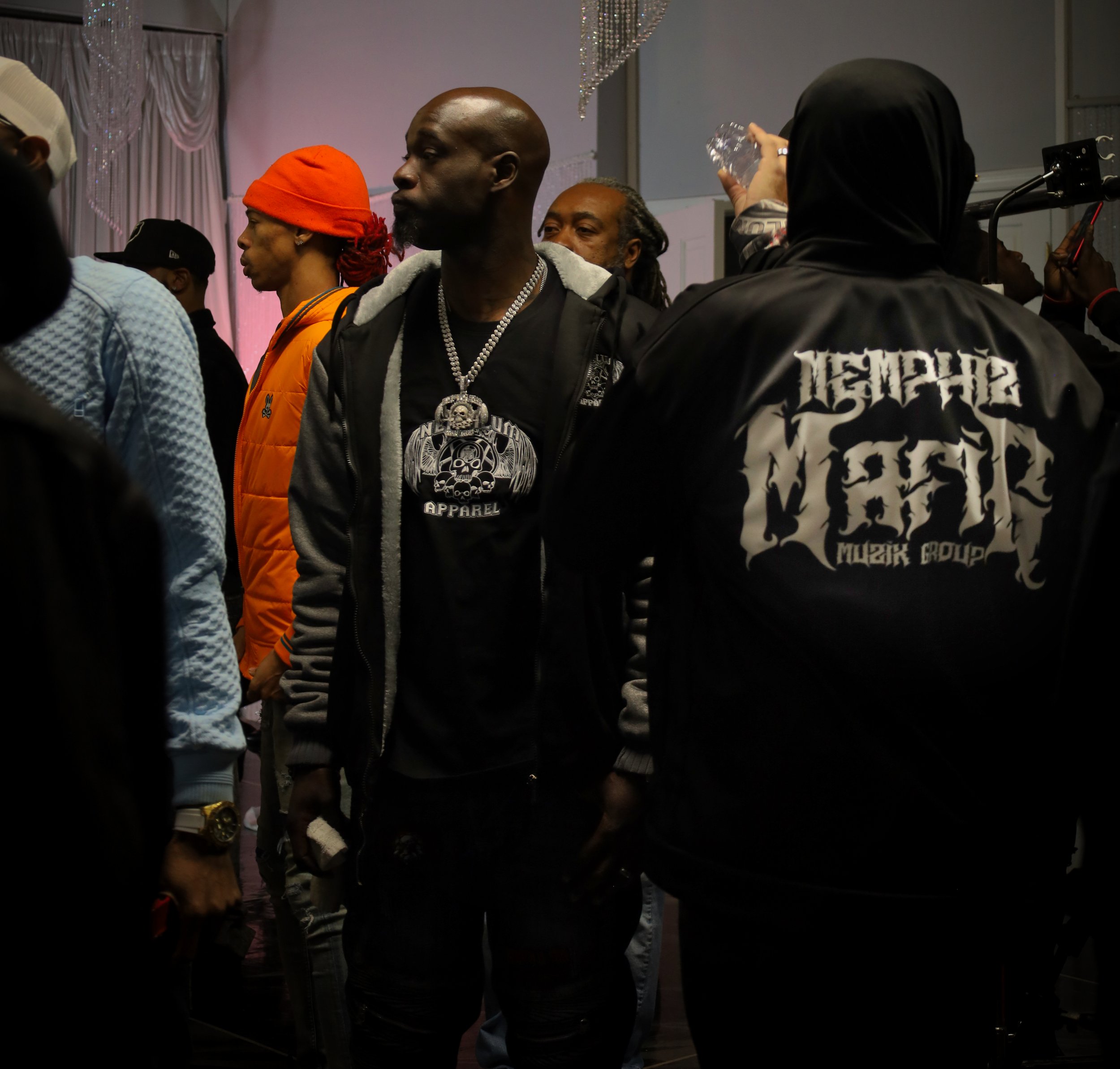 People standing indoors at an event, with some wearing casual and streetwear clothing, including hoodies, hats, and jewelry. One woman is drinking water from a plastic bottle, while others are engaged in conversations.