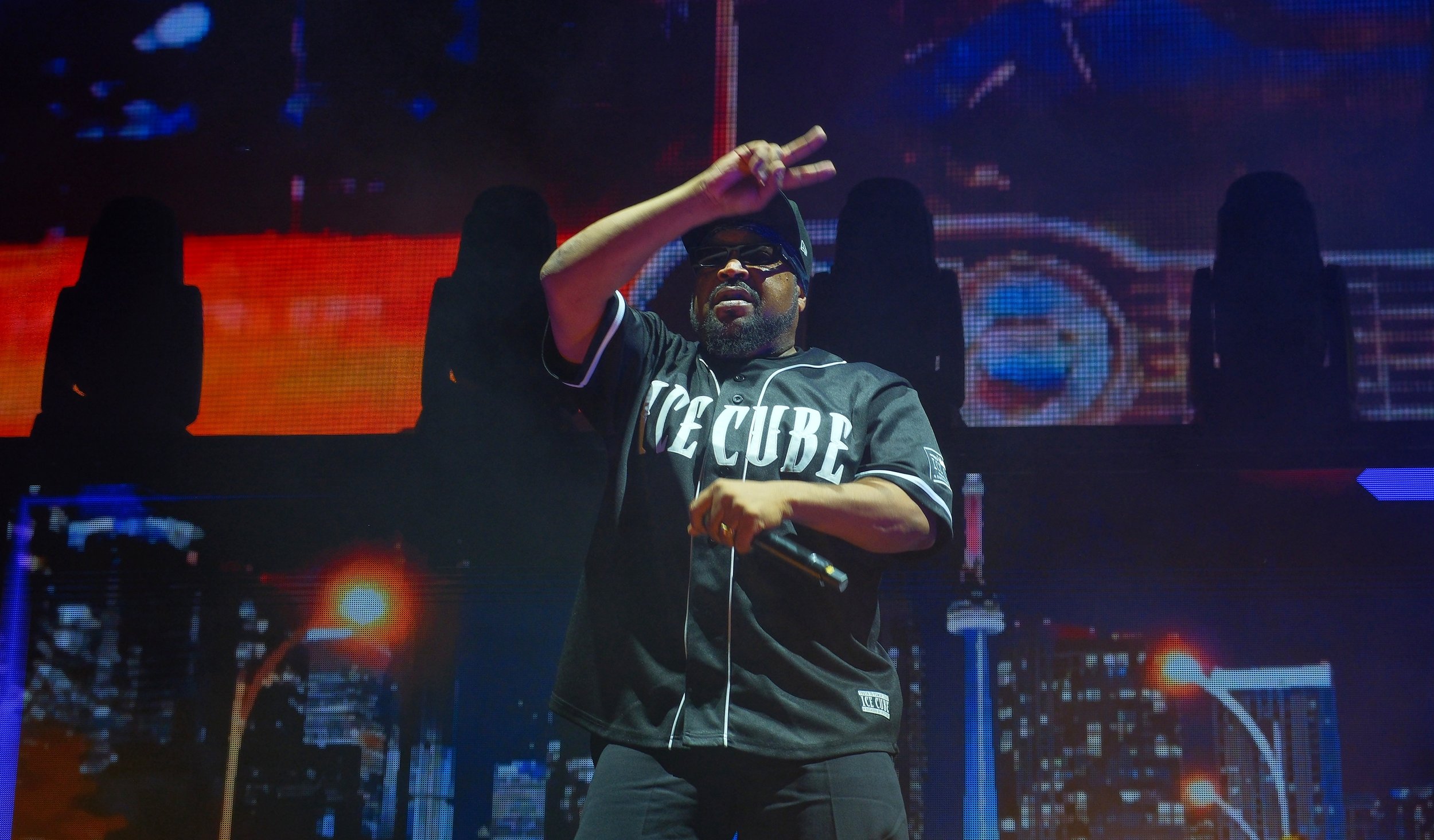 A performer wearing a black baseball jersey with the word 'ICECUBE' and black sunglasses is on stage making a peace sign with his fingers, with a vibrant digital background behind him.