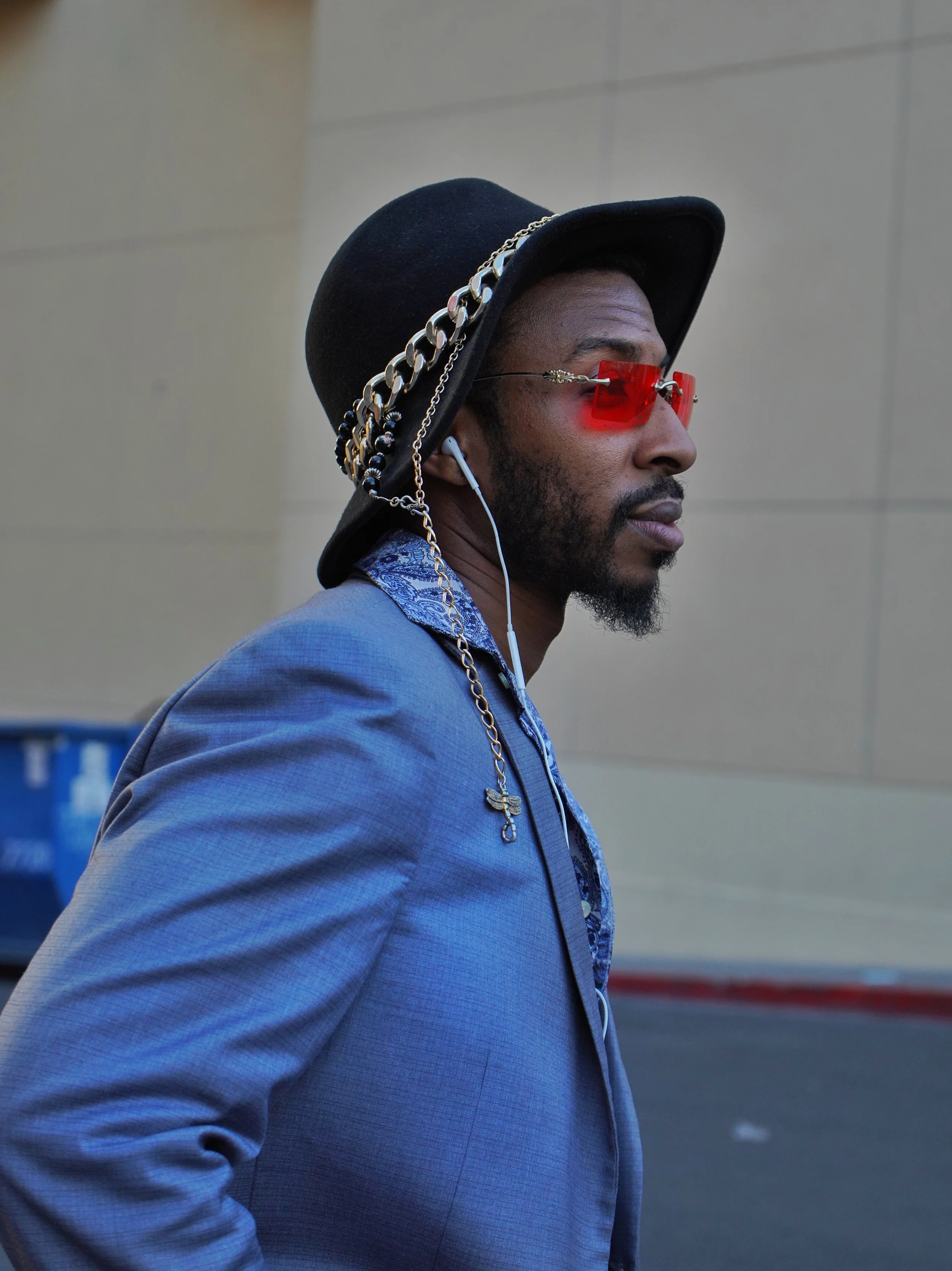 A man with dark skin wearing a black hat with metallic chains, red tinted sunglasses, a blue blazer, and earbuds, walking outdoors.