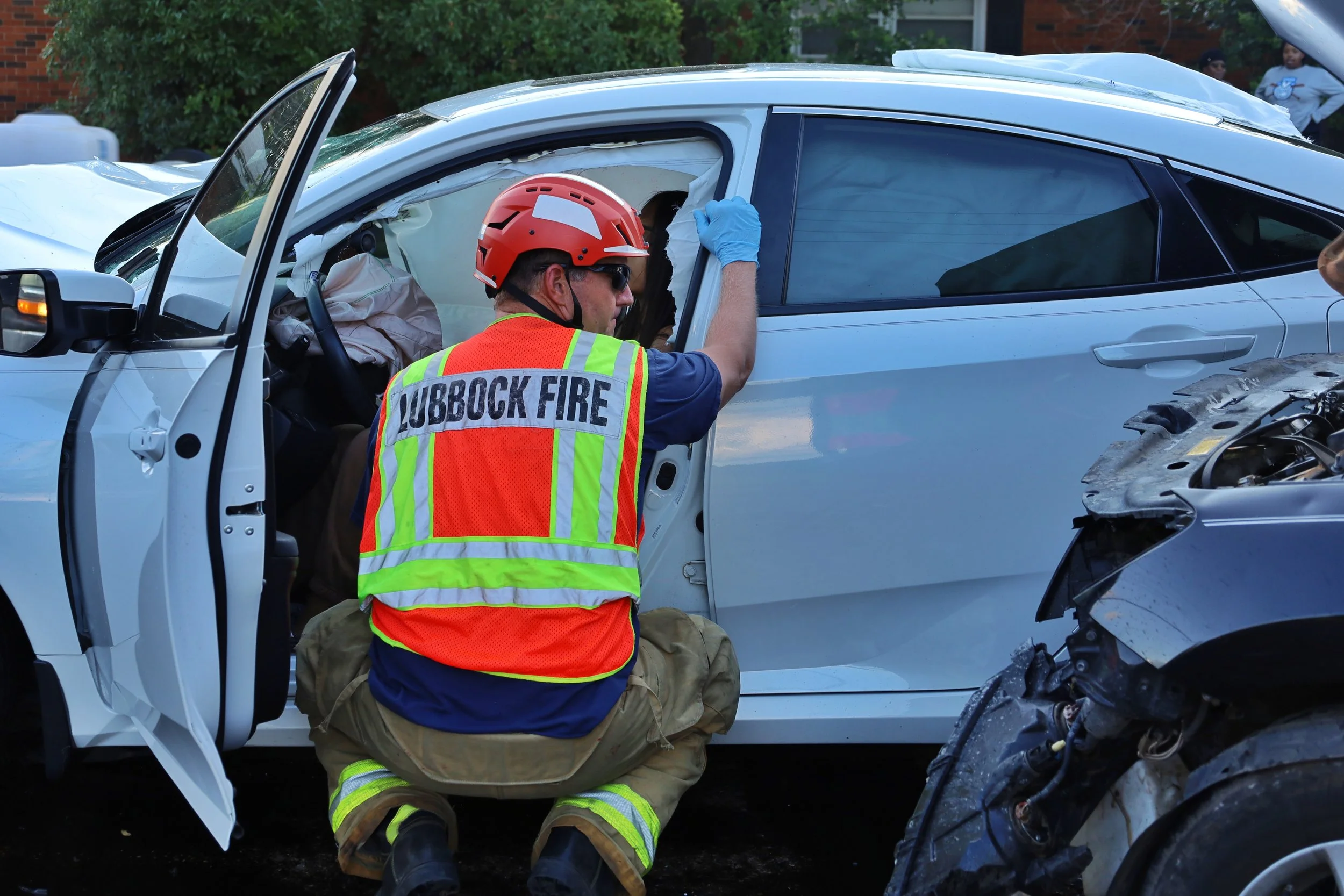 A firefighter from Lubbock Fire Department wearing a safety vest and helmet kneeling beside a damaged white car involved in a crash, inspecting the interior through the open driver’s side door.