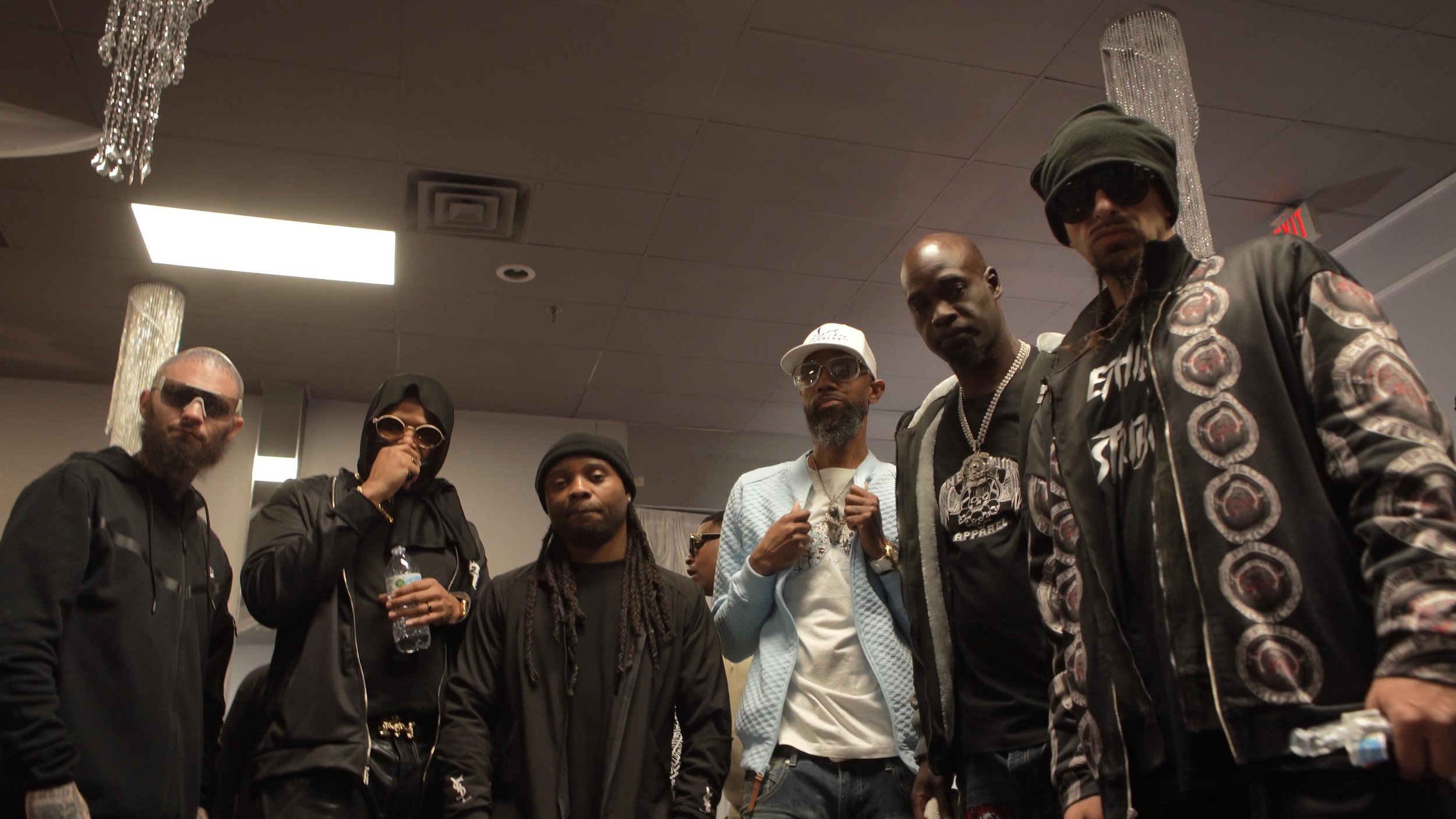 Group of six men posing indoors, some wearing sunglasses, hoodies, and jewelry, with a ceiling decorated with chandeliers in the background.