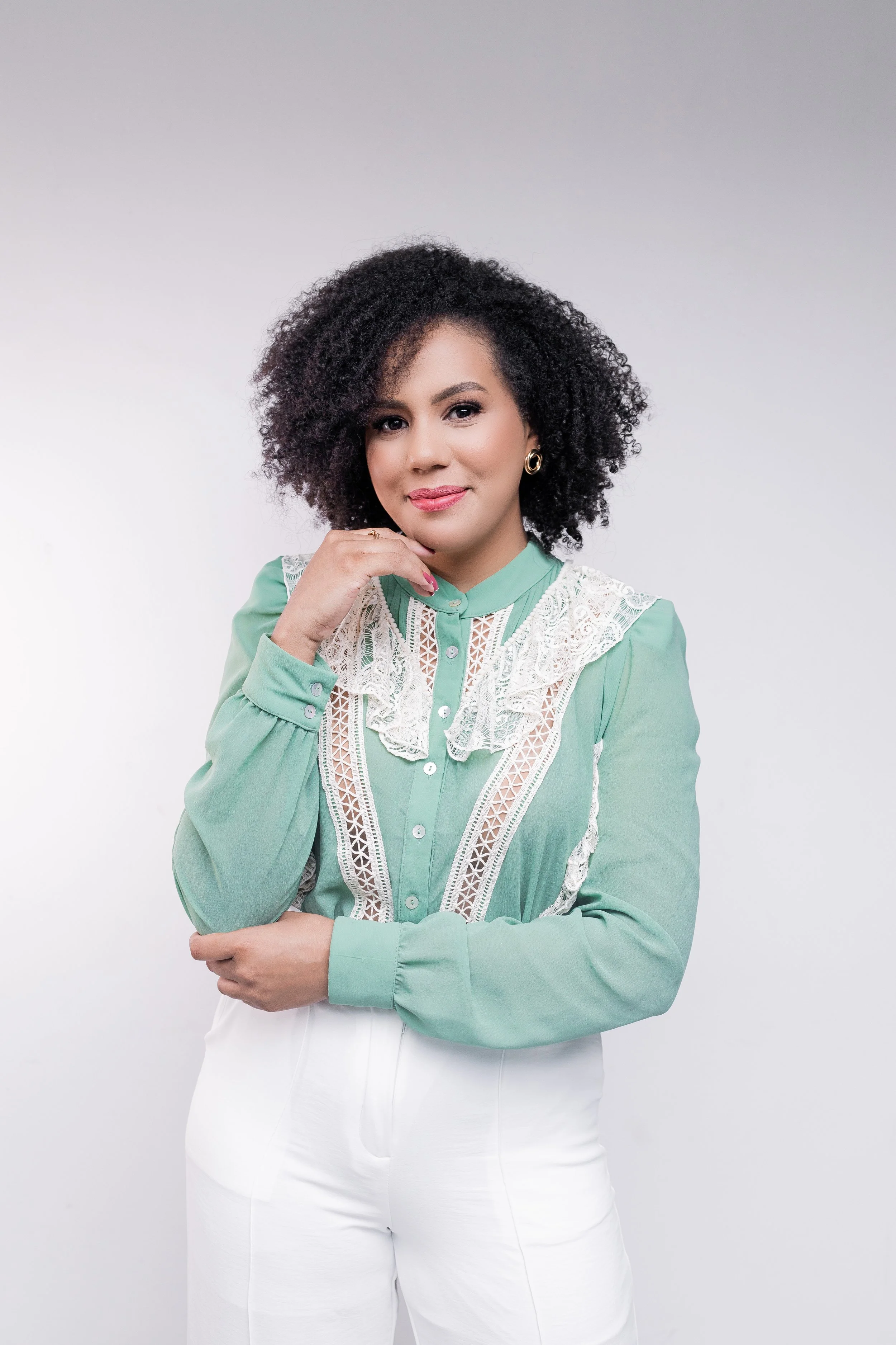 A woman with curly black hair wearing a mint green blouse with white lace details and white pants, standing against a plain white background.