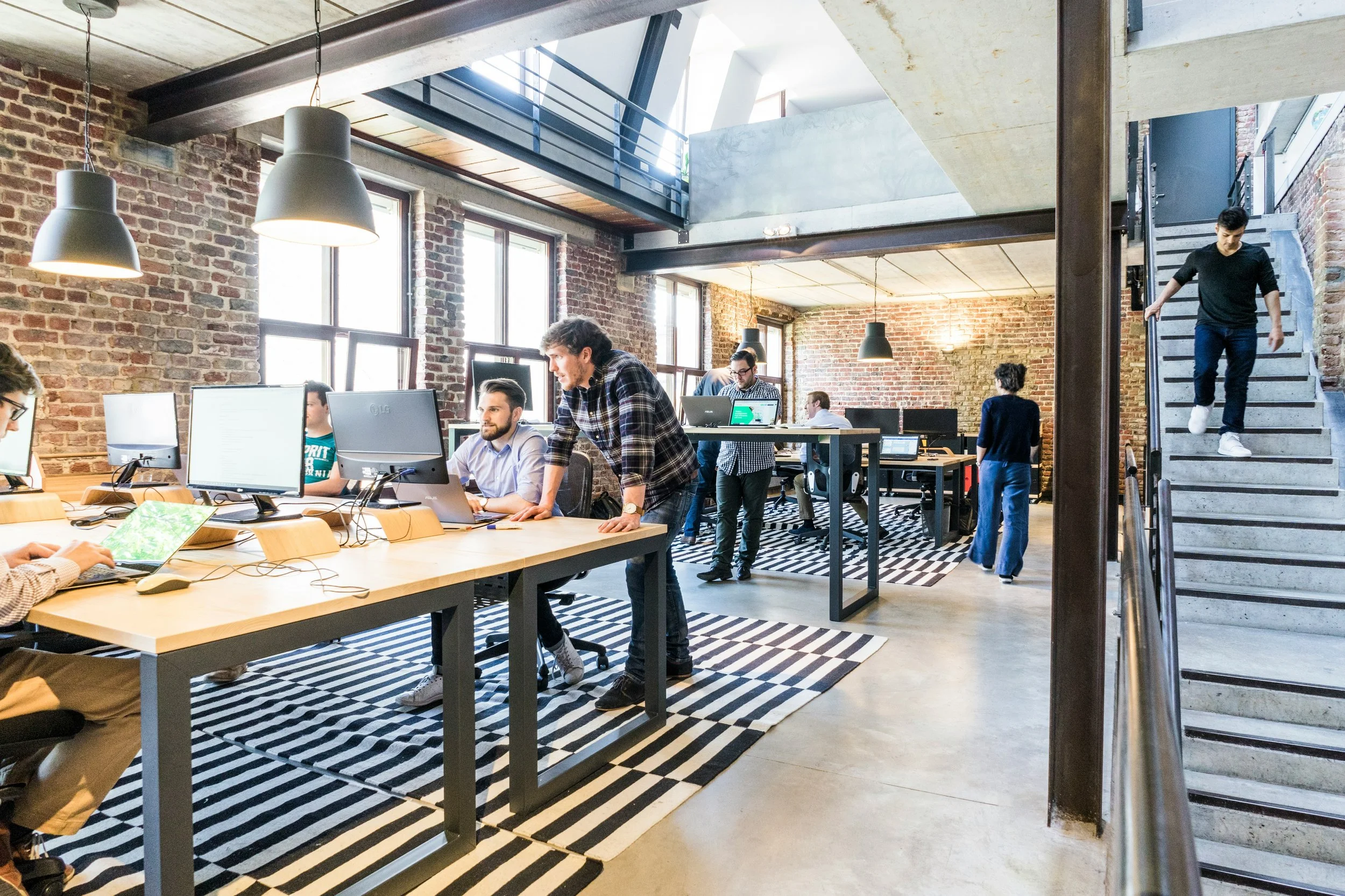 Modern open-plan office with exposed brick walls, large windows, and a mix of standing and seated employees working at desks with desktop computers. There are black pendant lights hanging from the ceiling, a striped area rug, and a staircase on the right side with a person walking down.