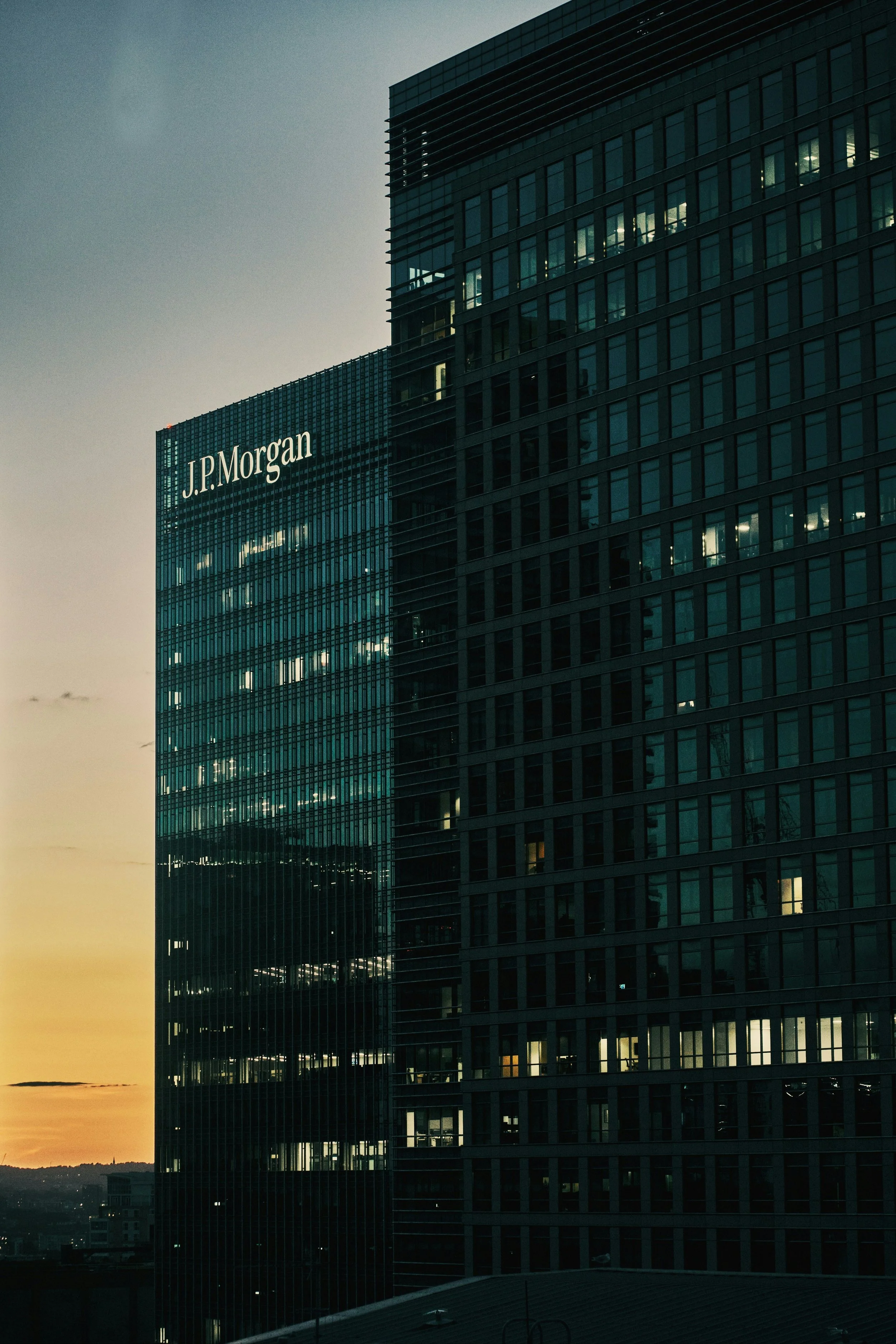 Skyline view of a modern office building with the J.P. Morgan logo, some office lights on inside, during sunset.