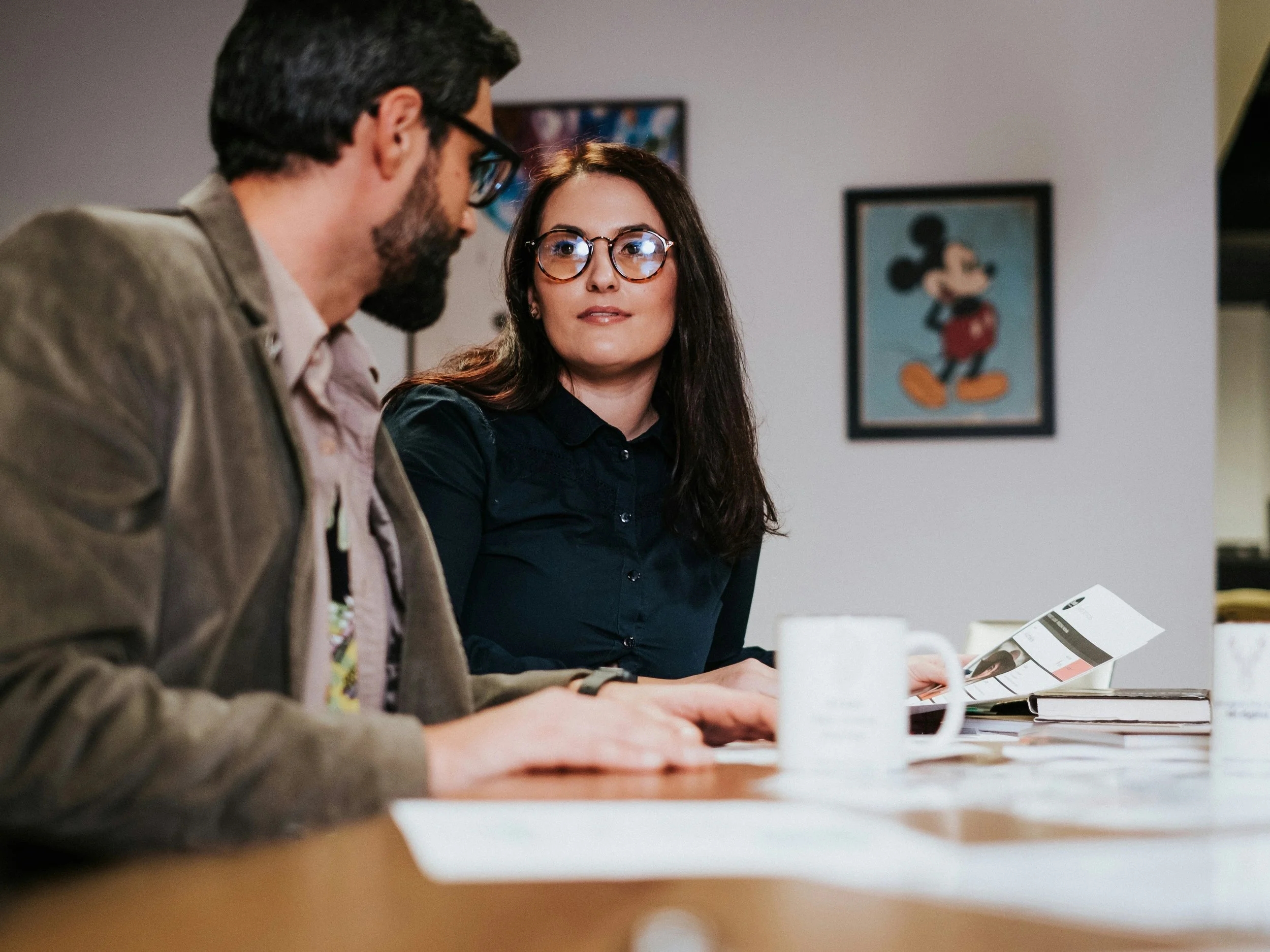 A man and a woman sit at a table in a meeting room, with the woman holding a brochure. The woman looks at the man who is looking down at the table. Behind them, there are framed pictures on the wall, including a Mickey Mouse illustration.
