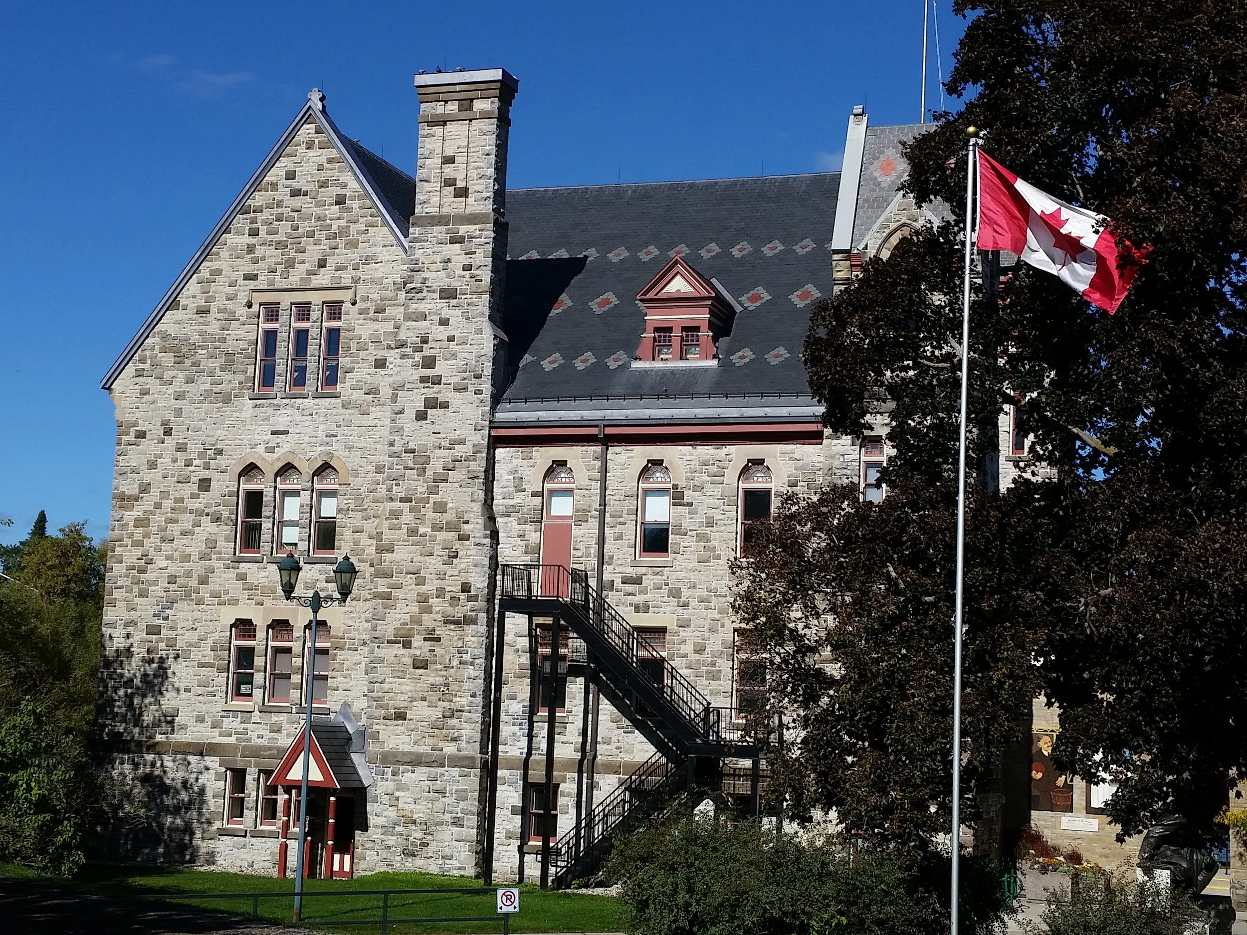 A historic stone building with red window frames and a staircase on the outside, partially obscured by a large tree, with the Canadian flag on a flagpole, under a clear blue sky.