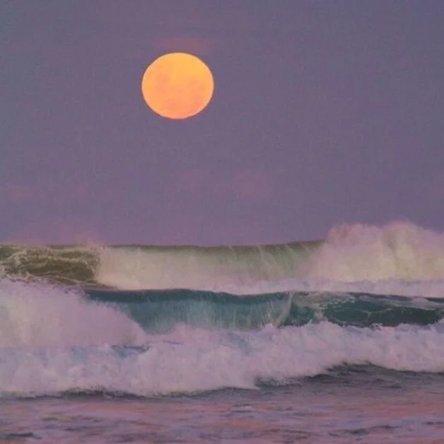 Over the ocean at sunset with a large, orange moon in the sky and waves crashing on the shore.