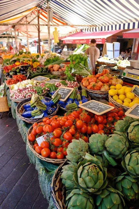 Fresh vegetables and produce displayed in baskets at an outdoor farmer's market stall, including tomatoes, artichokes, onions, lemons, and eggplants, with a striped canopy overhead.
