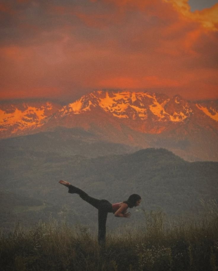 A person practicing yoga outdoors on a grassy field with mountains in the background at sunset.