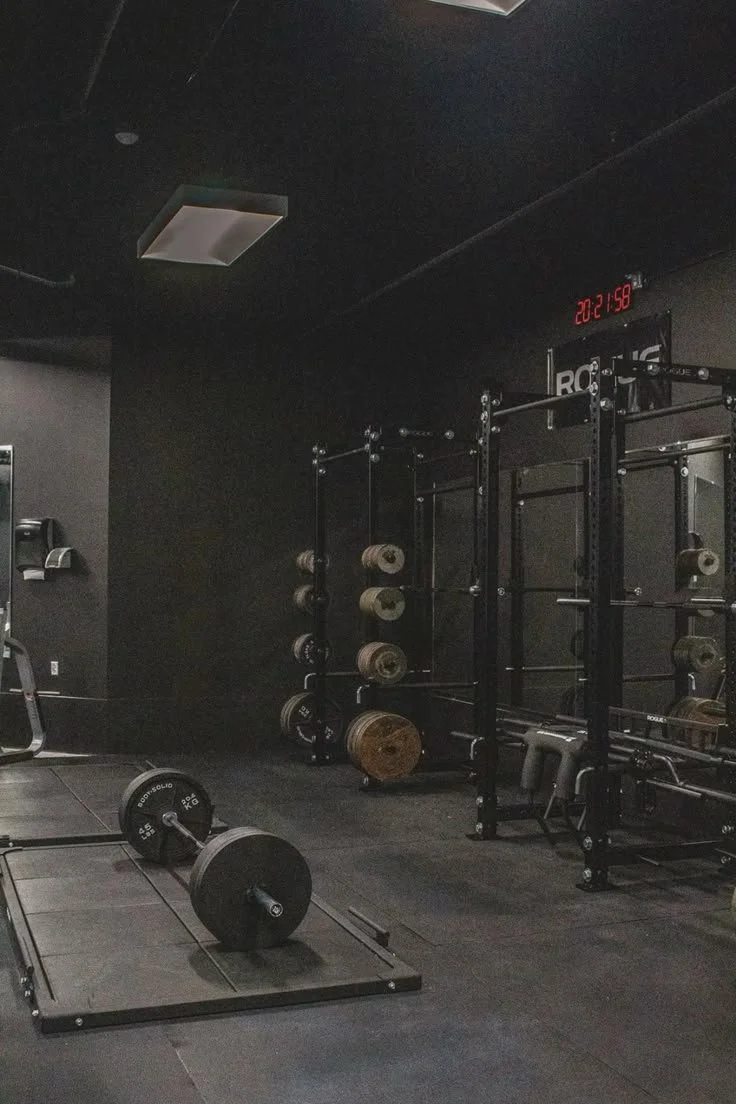 Empty gym with weightlifting equipment including a barbell on the floor, a rack of various weights on wall-mounted shelves, and a clock showing 8:21 PM.