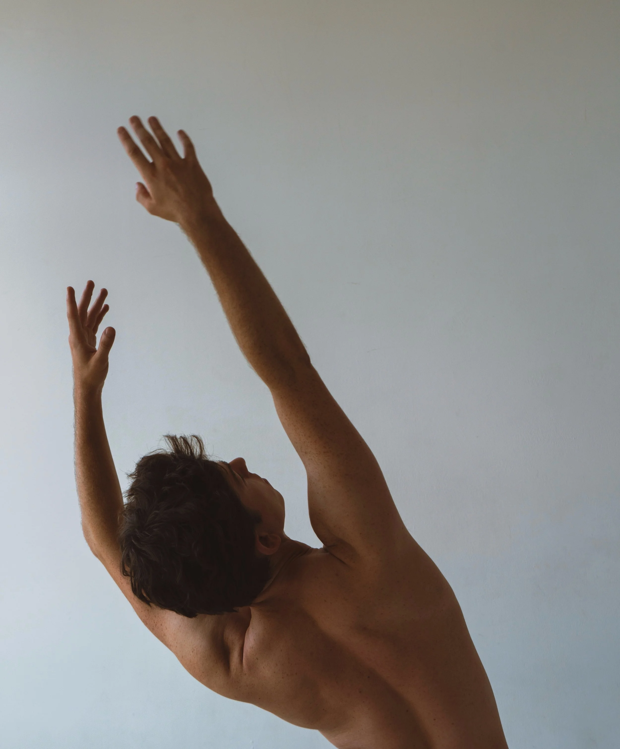 Back view of a shirtless ballet dancer with short dark hair, raising their arms above their head, against a plain light-colored wall. The dancer is showing the strentgh and form of ballet and pilates. Cian Hughes, dancer and pilates instructor