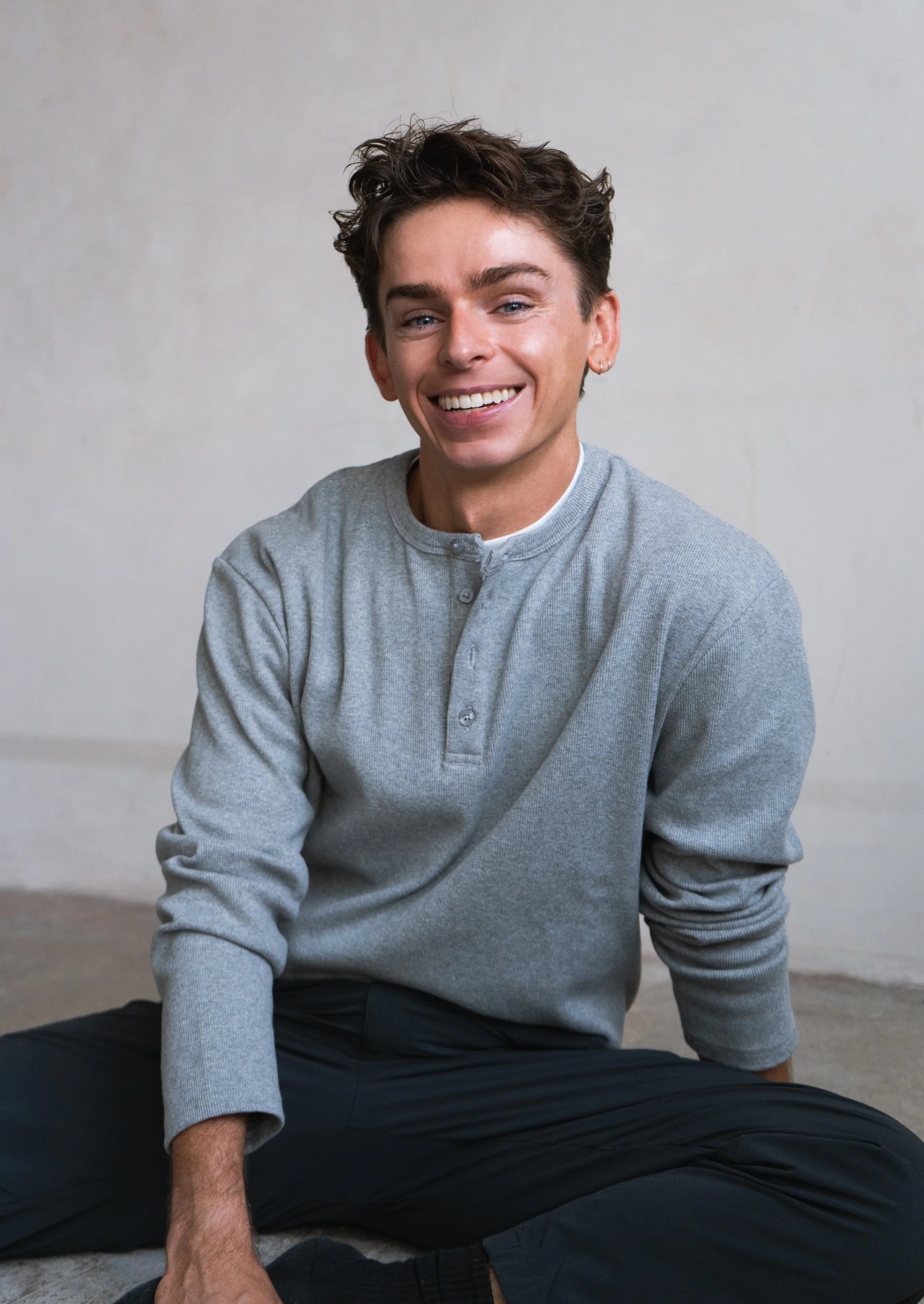 A young man with short, dark, curly hair smiling while sitting against a plain, light-colored wall, dressed in a light gray, long-sleeved, button-up shirt and dark pants. Cian Hughes, founder of Poem Pilates.