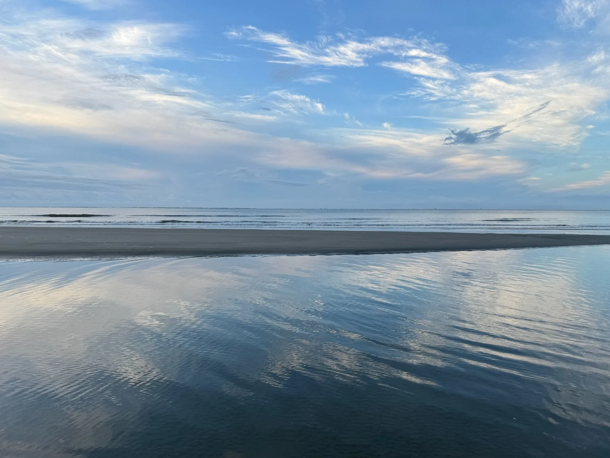 Calm ocean with gentle waves and cloudy sky reflected on water surface, viewed from the shoreline.