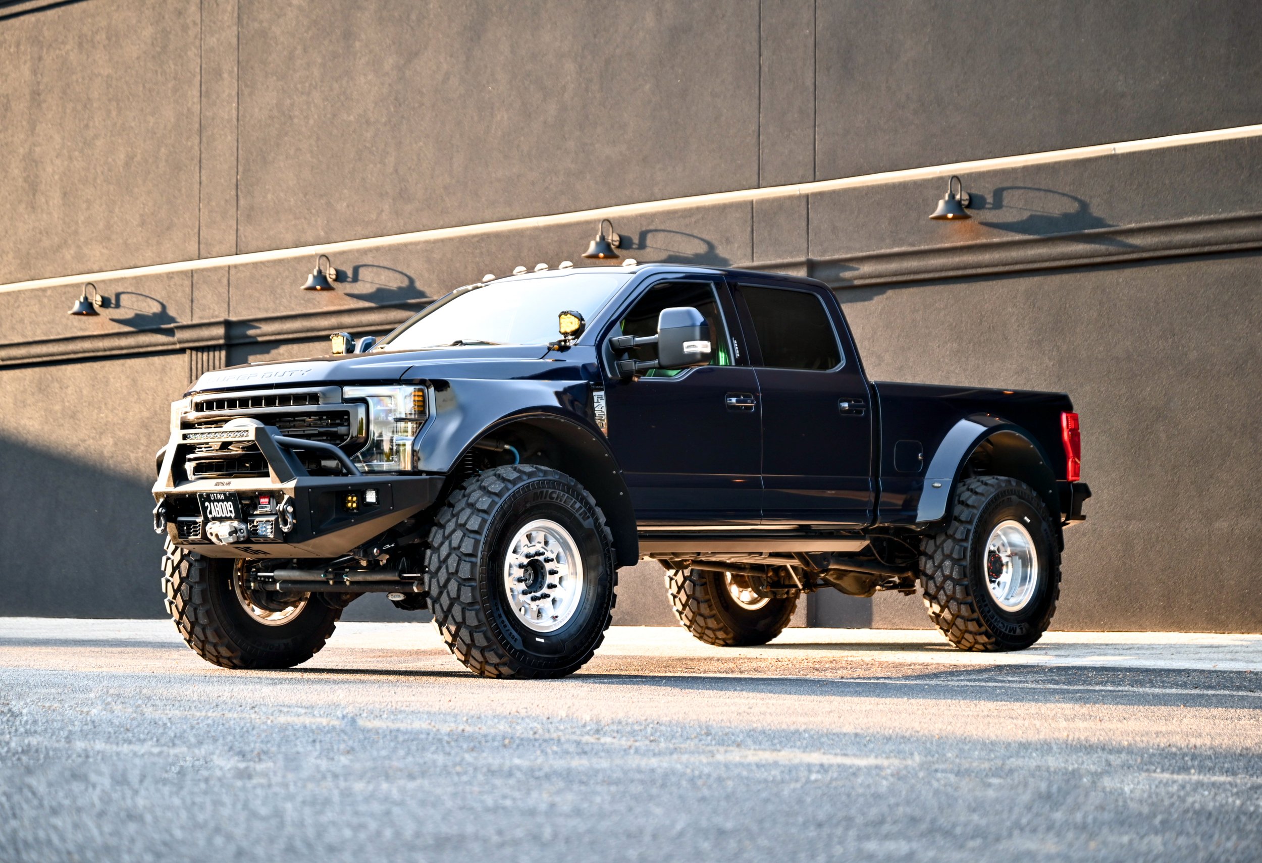 A black and grey pickup truck with rugged tires parked on a concrete surface in front of a textured grey wall.