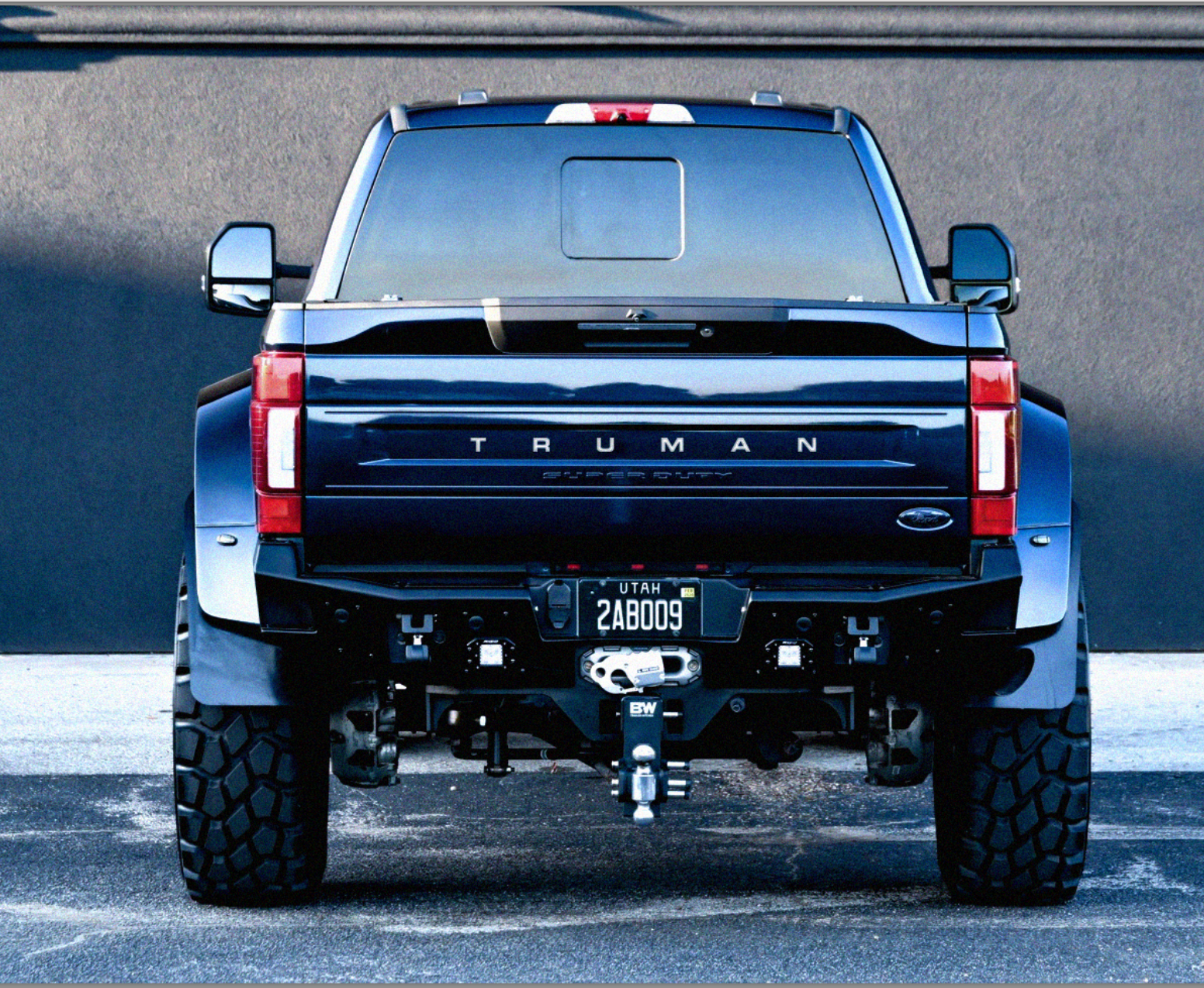 Rear view of a black Ford pickup truck with large off-road tires, Maryland license plate, and a tow hitch, parked on pavement against a dark wall.
