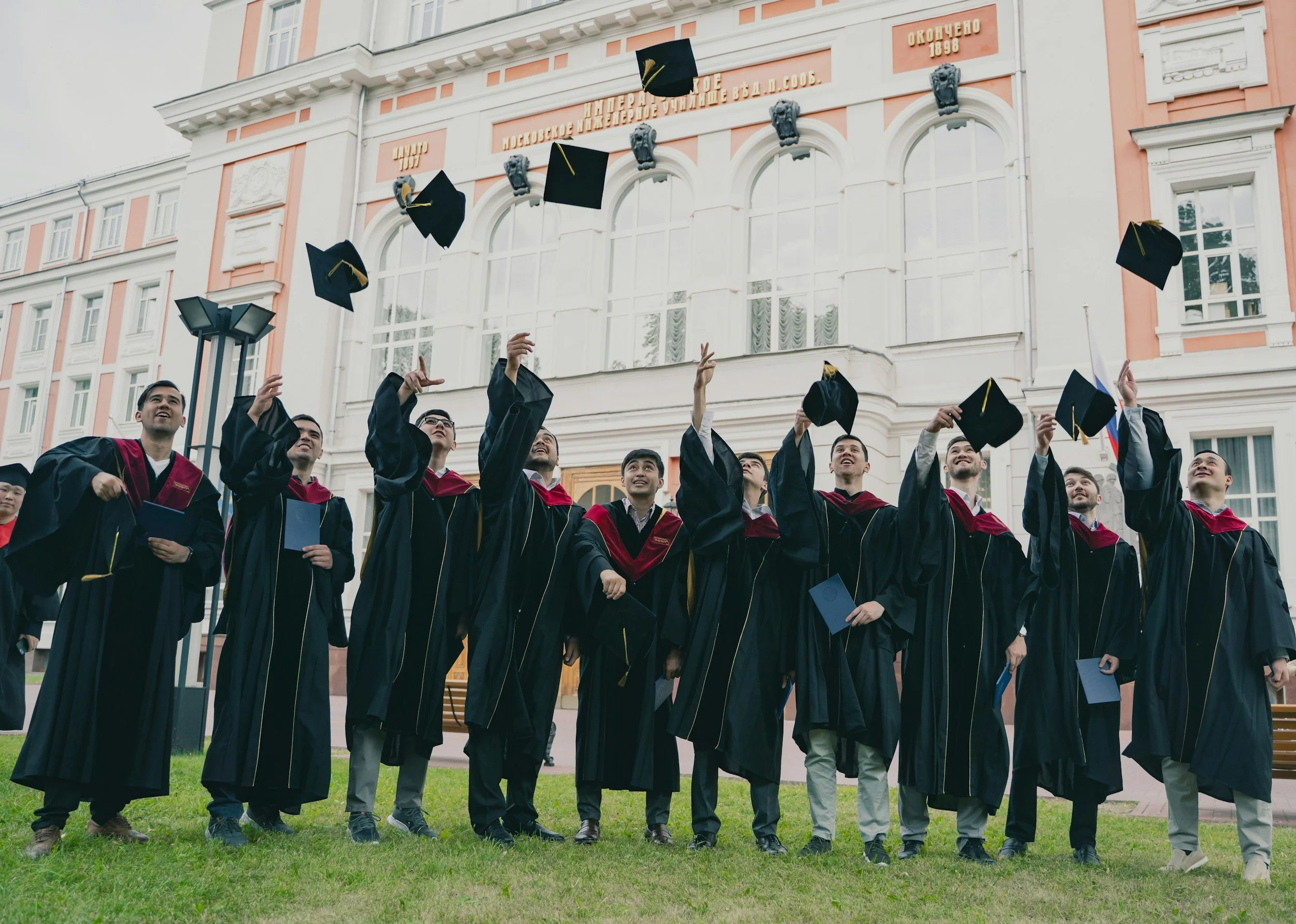Group of graduates in cap and gown celebrating on campus lawn during graduation ceremony, tossing caps into the air in front of university building.