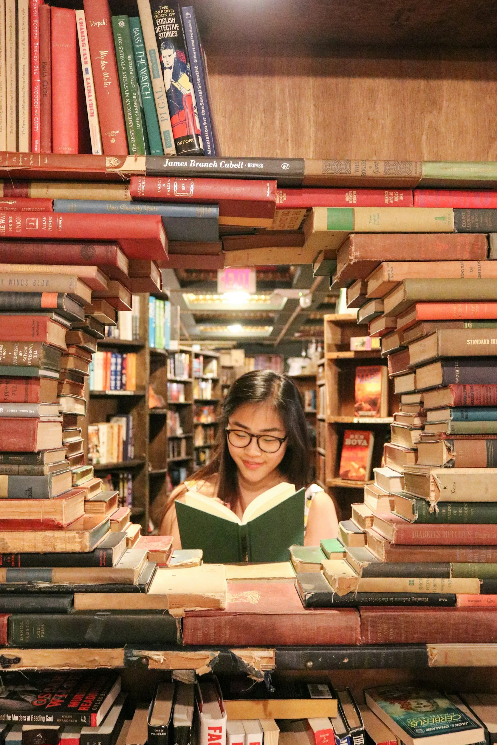 A woman with glasses reading a book inside a bookstore, framed by stacks of books forming an archway.